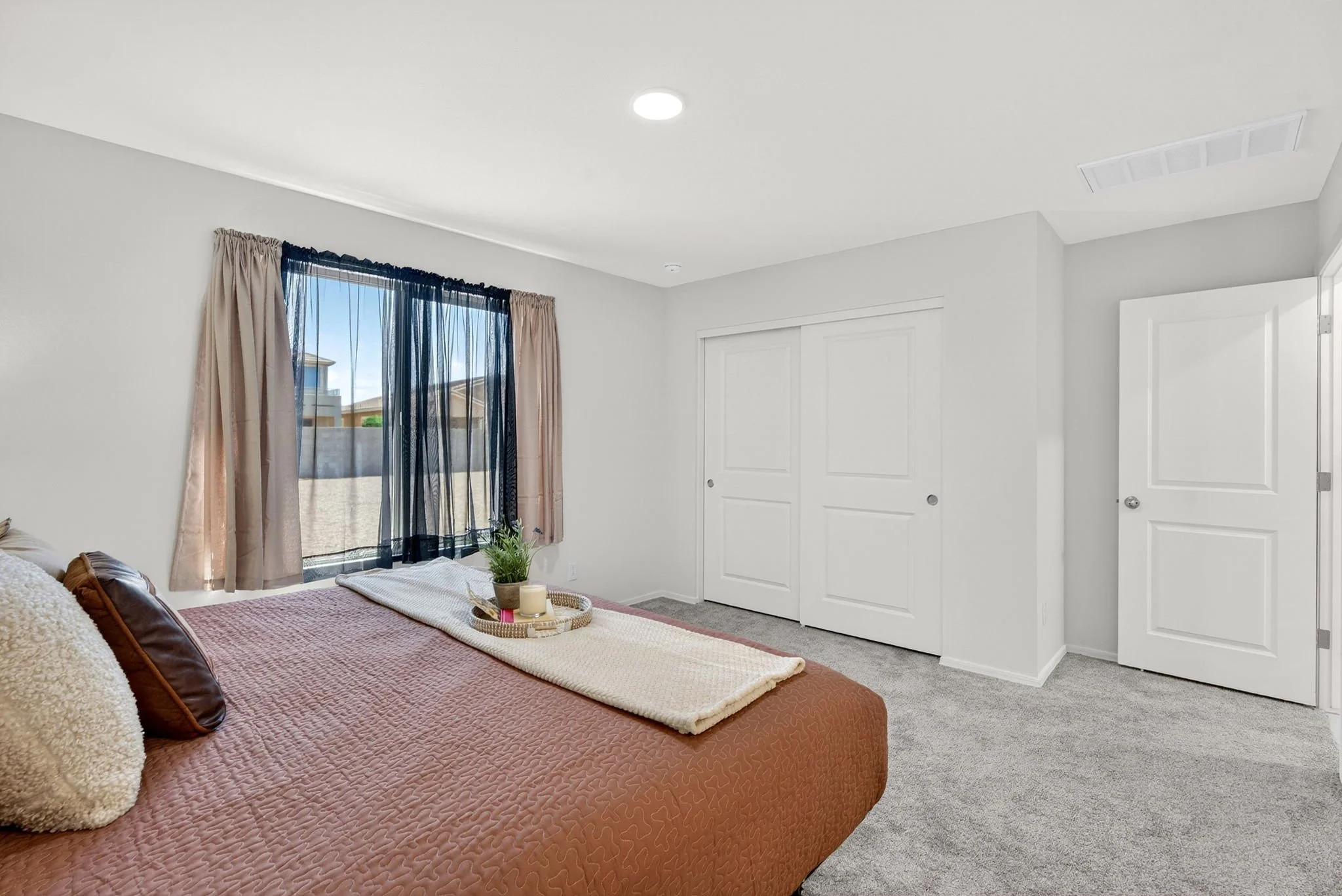 A bedroom with a window with sheer black curtains and beige drapes, a bed with a pink quilt, pillows, and a wooden tray with a plant and candles. There are two closed white closet doors and a white door to the room, with light gray walls and gray car