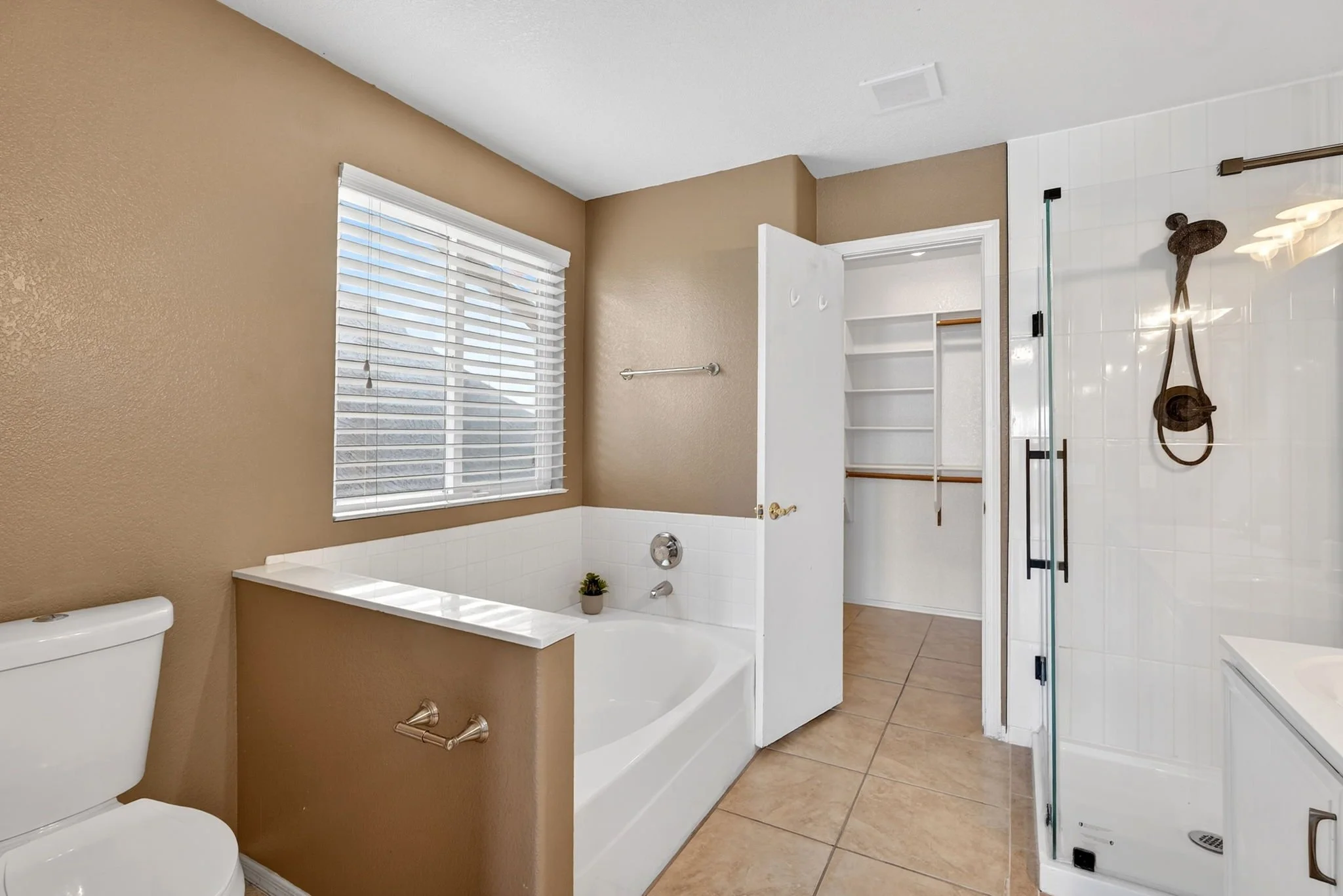 Bathroom with beige walls, a white toilet, a bathtub with a potted plant, a window with blinds, a glass shower enclosure, and a walk-in closet with shelves.