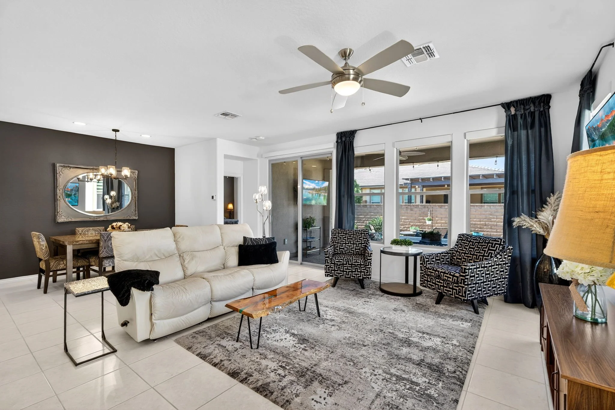 Spacious living room with white tiled floor, white and dark gray walls, ceiling fan, large windows with dark curtains, beige sofa, patterned armchairs, and various lamps and decorations.
