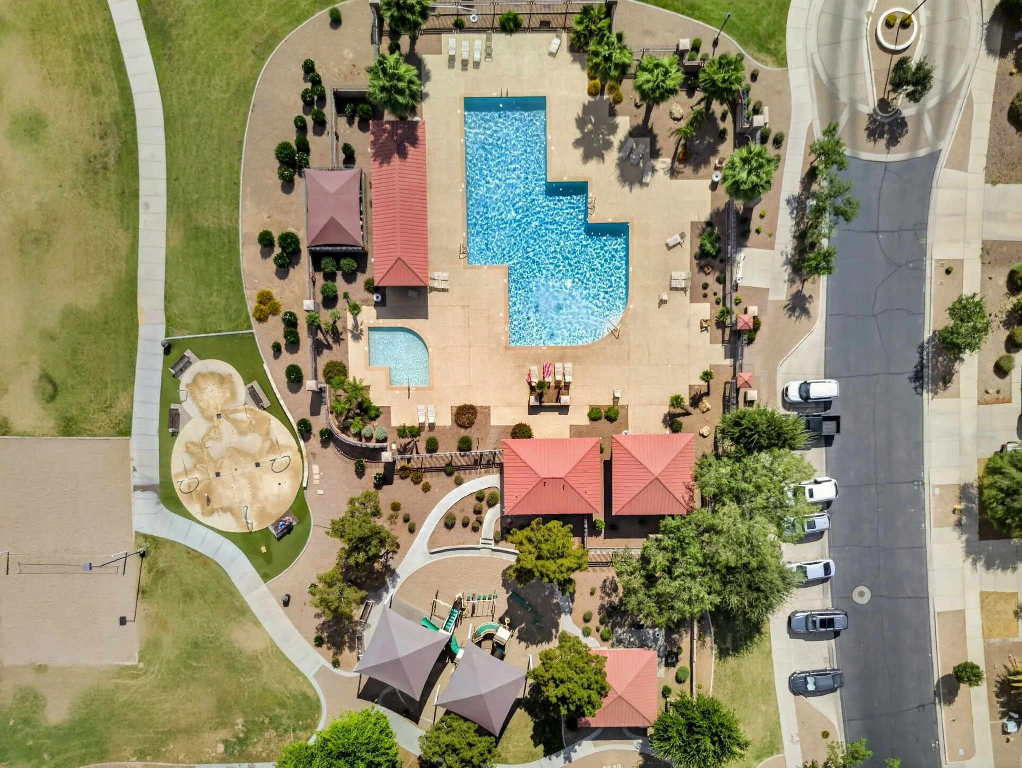 Aerial view of a community park with a swimming pool, surrounding lounge chairs, shaded structures, playground, walking paths, trees, and parked cars.