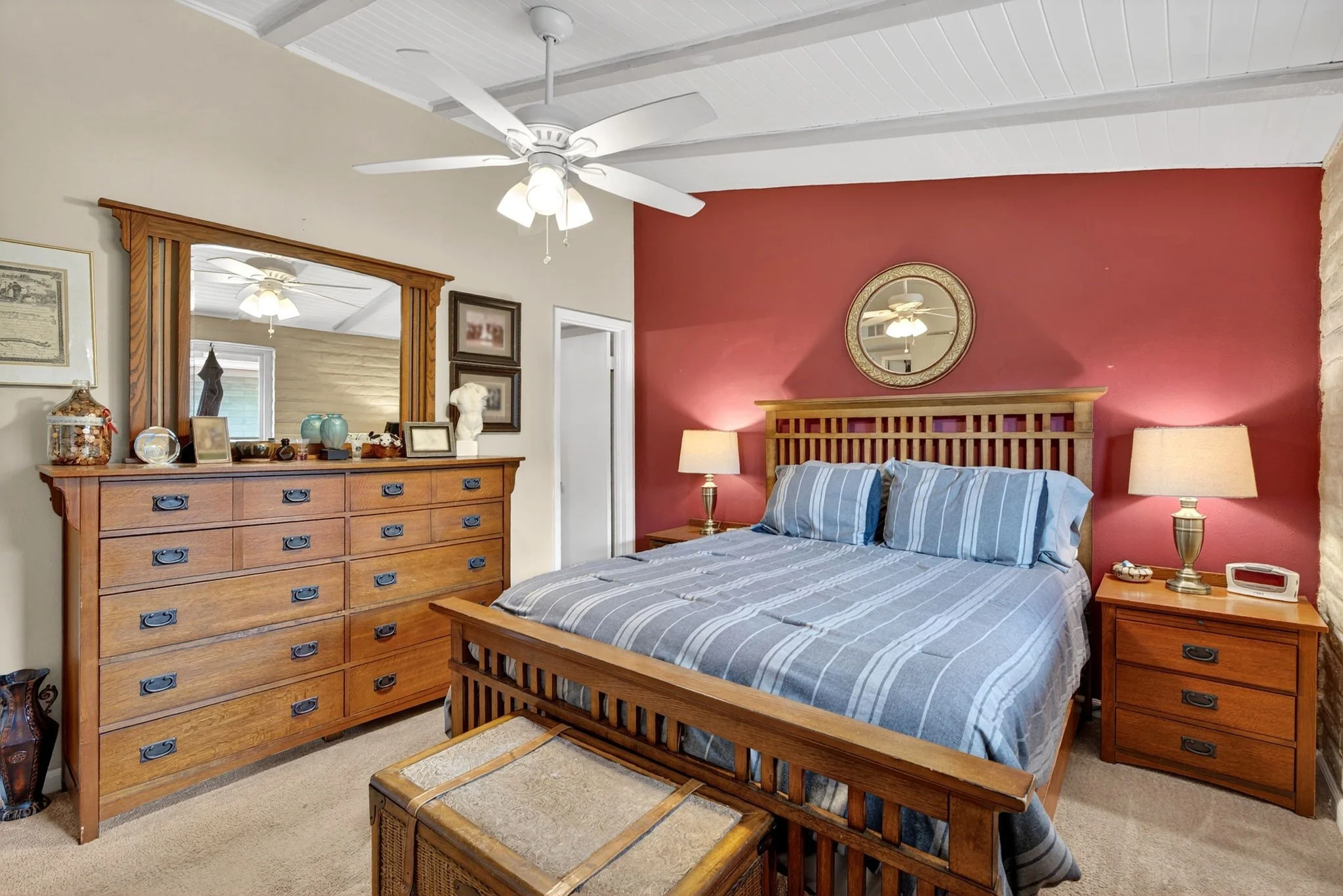A bedroom with a bed that has blue and white striped bedding, a wooden headboard, and matching side tables with lamps. There is a wooden dresser with various decorations and a mirror on the wall above. The room features a white ceiling with a ceiling