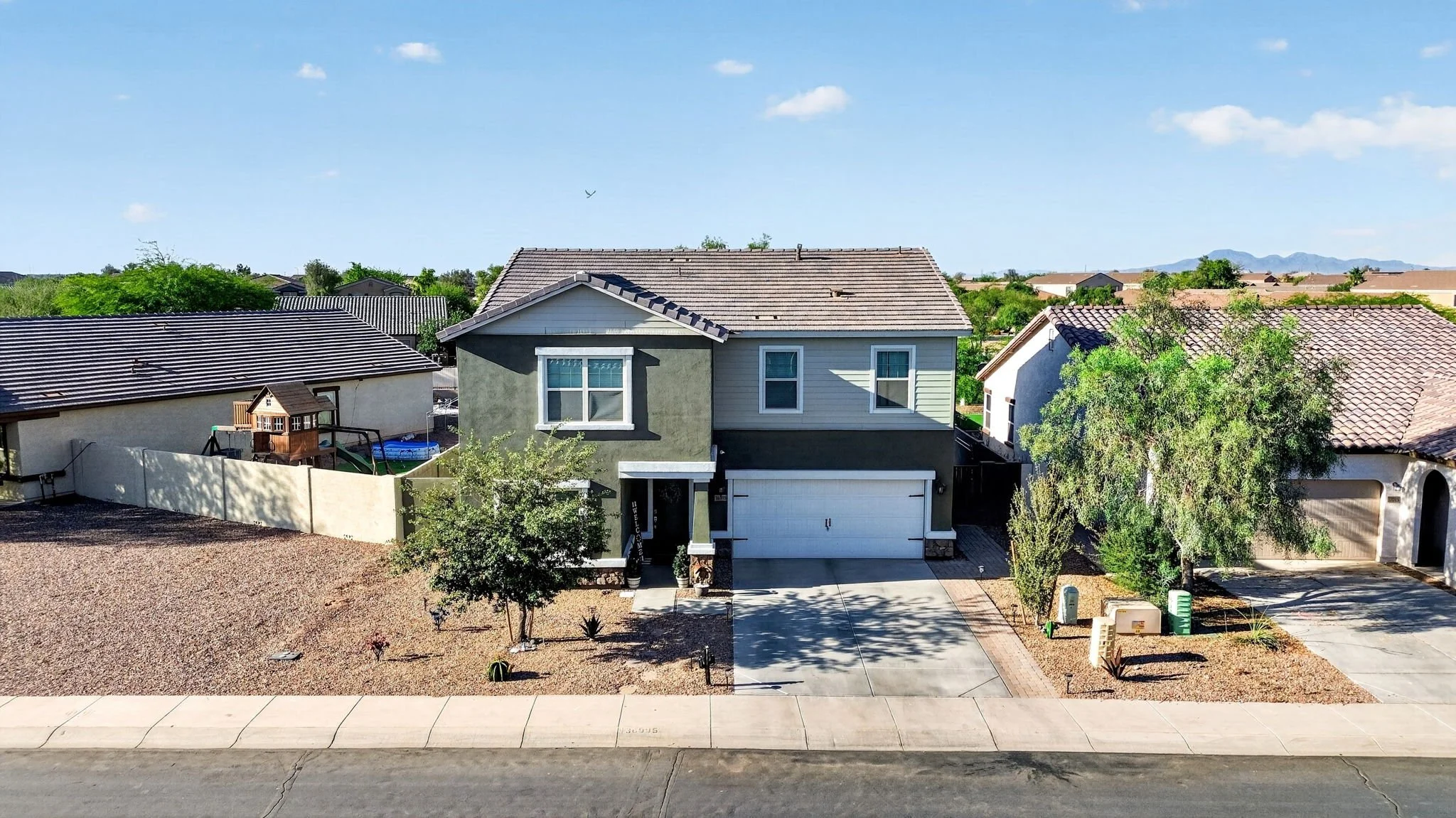 A two-story suburban house with a gray exterior, white garage door, and a sloped roof. The front yard features a tree, desert landscaping, and a driveway. Neighboring houses and distant mountains are visible under a blue sky.