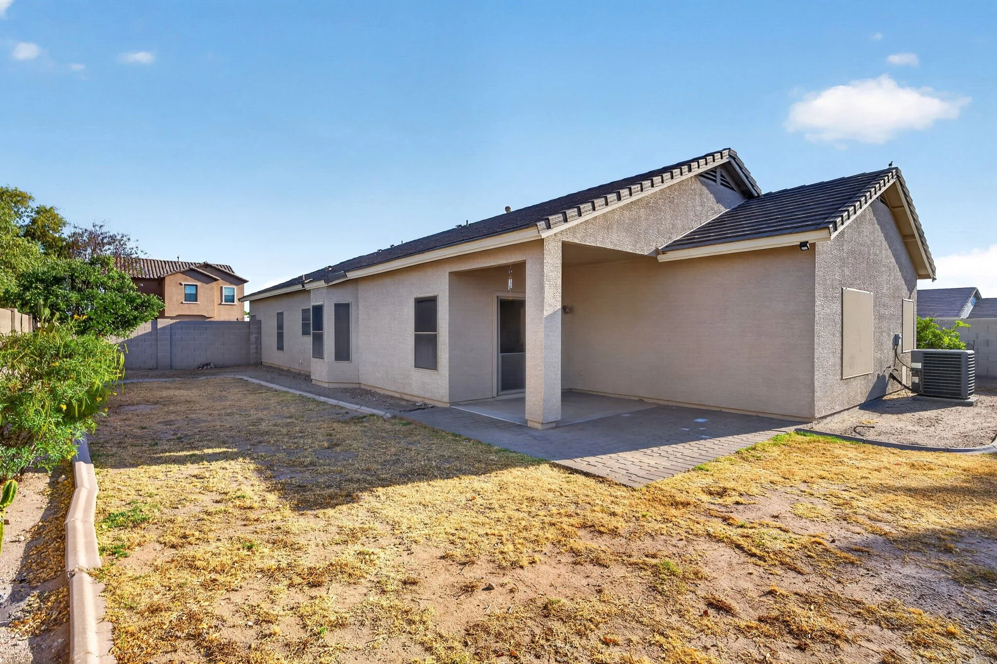 Backyard of a house with beige stucco walls, a concrete patio, and an air conditioning unit. The yard has patchy grass and dirt, a small garden bed on the left with plants and a tree, and a brick pathway leading to the house. A neighboring house with