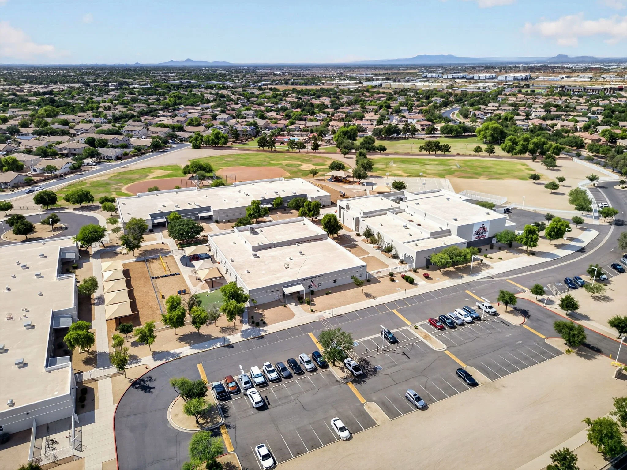Aerial view of a suburban area with houses, green trees, parking lots, and sports fields, with distant mountains in the background.