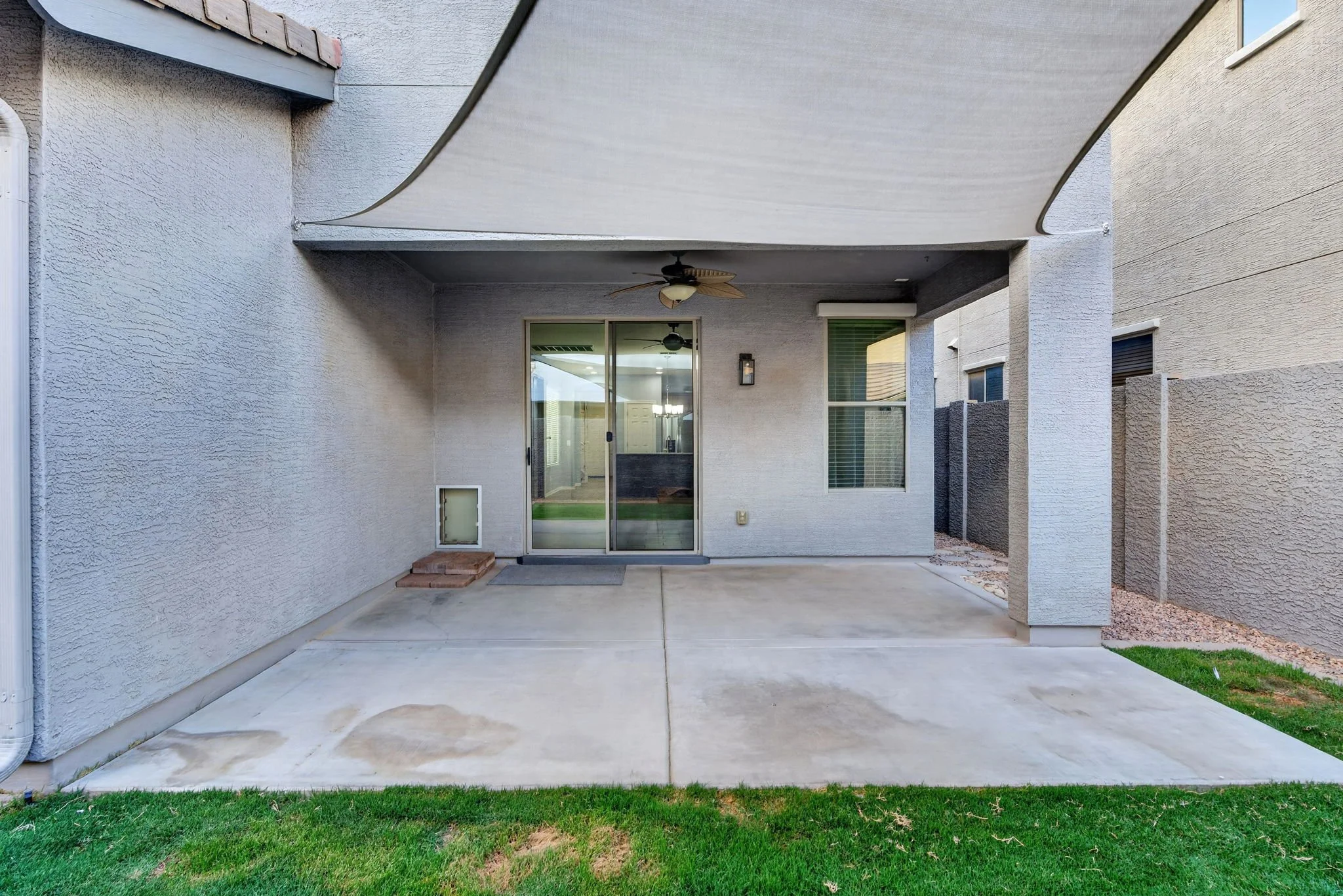 Backyard patio area with a sliding glass door, ceiling fan, and a stone step at the doorway, surrounded by beige stucco walls and a small grassy area.