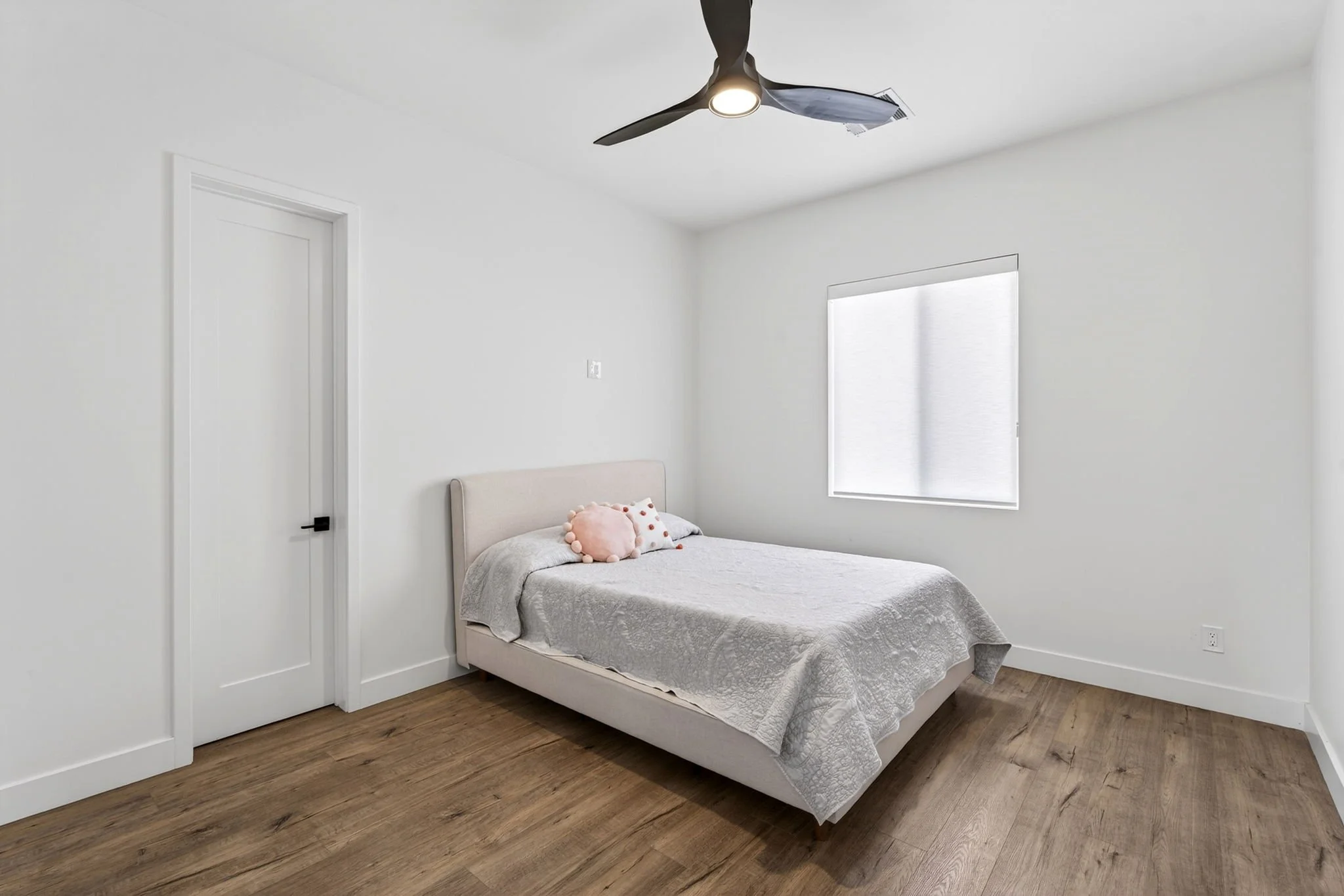 A minimalist bedroom with white walls and ceiling. There is a white door on the left and a window with closed blinds on the right. A bed with a beige upholstered headboard, dressed in a light gray quilted bedspread, is positioned against the wall. On
