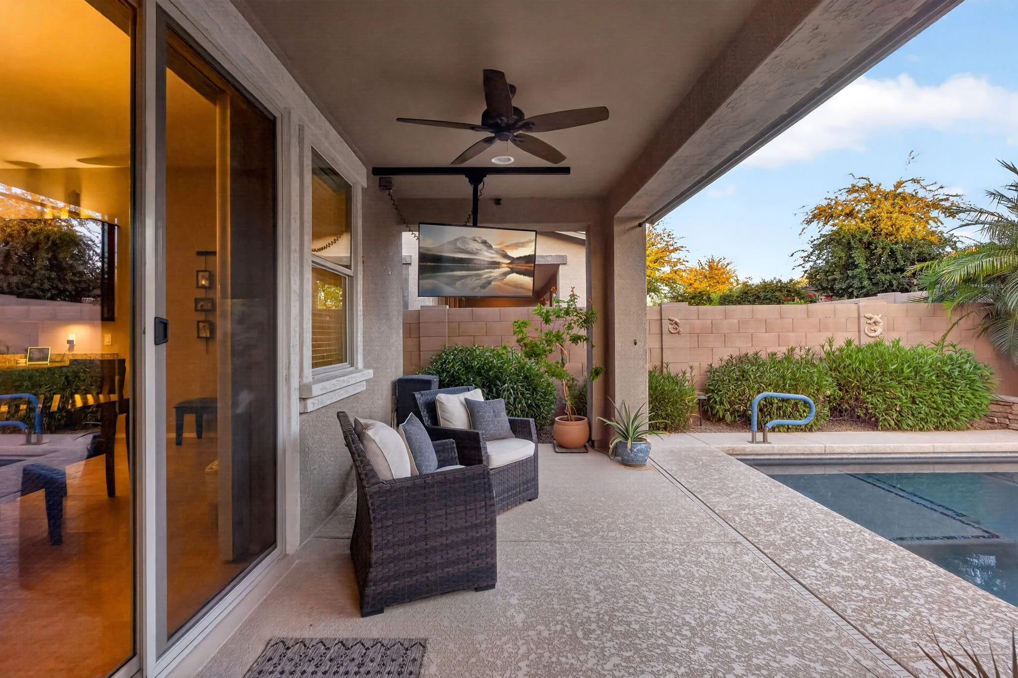 Covered patio area with outdoor seating, potted plants, ceiling fan, and view of trees and fence beyond the pool.