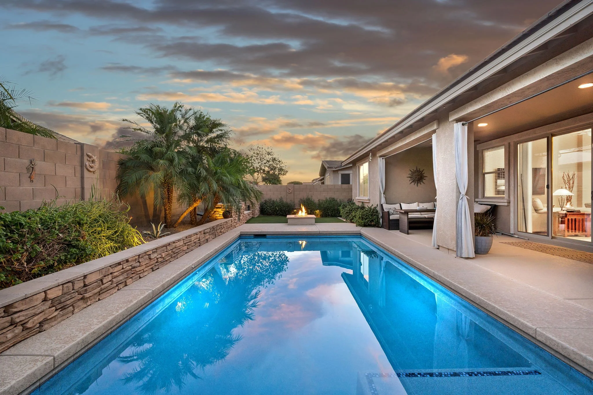 Backyard at dusk with a swimming pool, landscaped garden, firepit, and a covered patio area with furniture, under a partly cloudy sky.