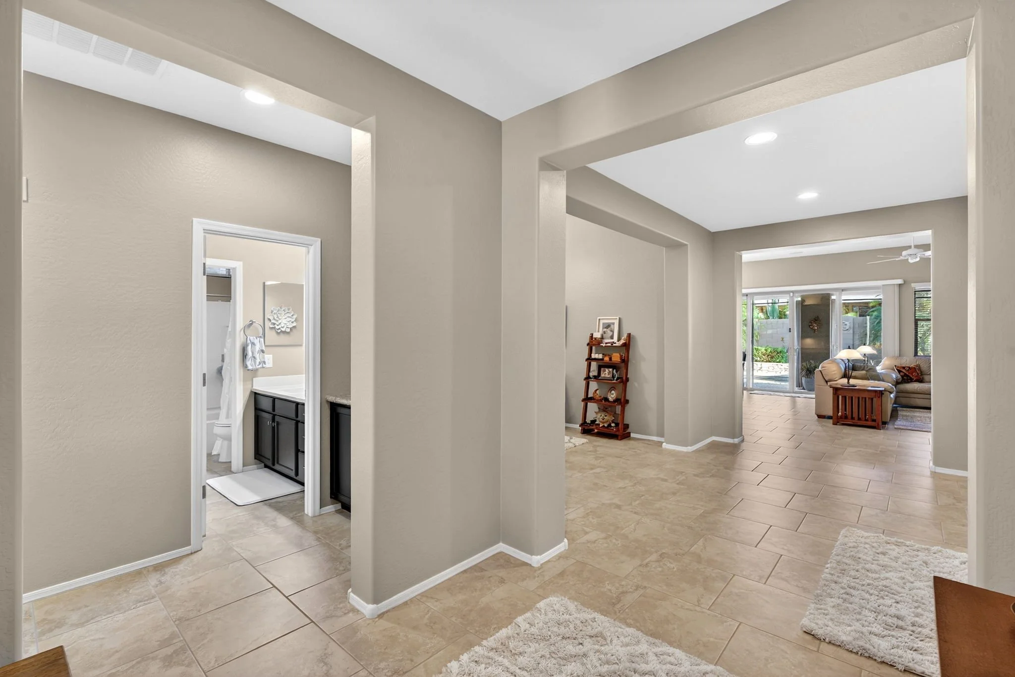Interior view of a home with beige walls, tiled flooring, and open living spaces, including a living room with beige sofas and large windows showing outdoor greenery.