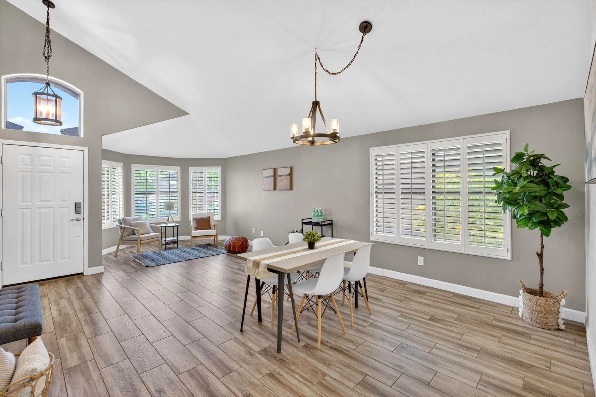 A spacious living room and dining area with large windows and white window shutters, modern chandelier, wooden flooring, and various chairs and tables with decor.