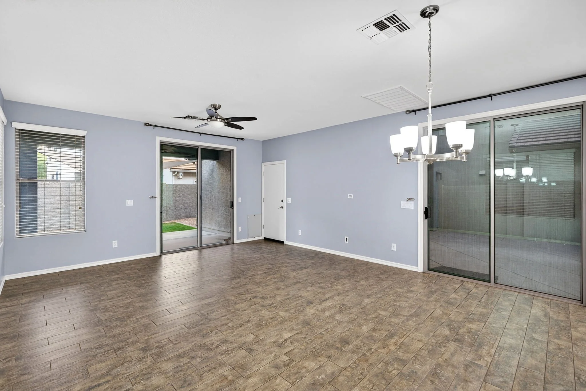 Empty living room with light blue walls, hardwood flooring, ceiling fan, chandelier, sliding glass doors, and windows with blinds.