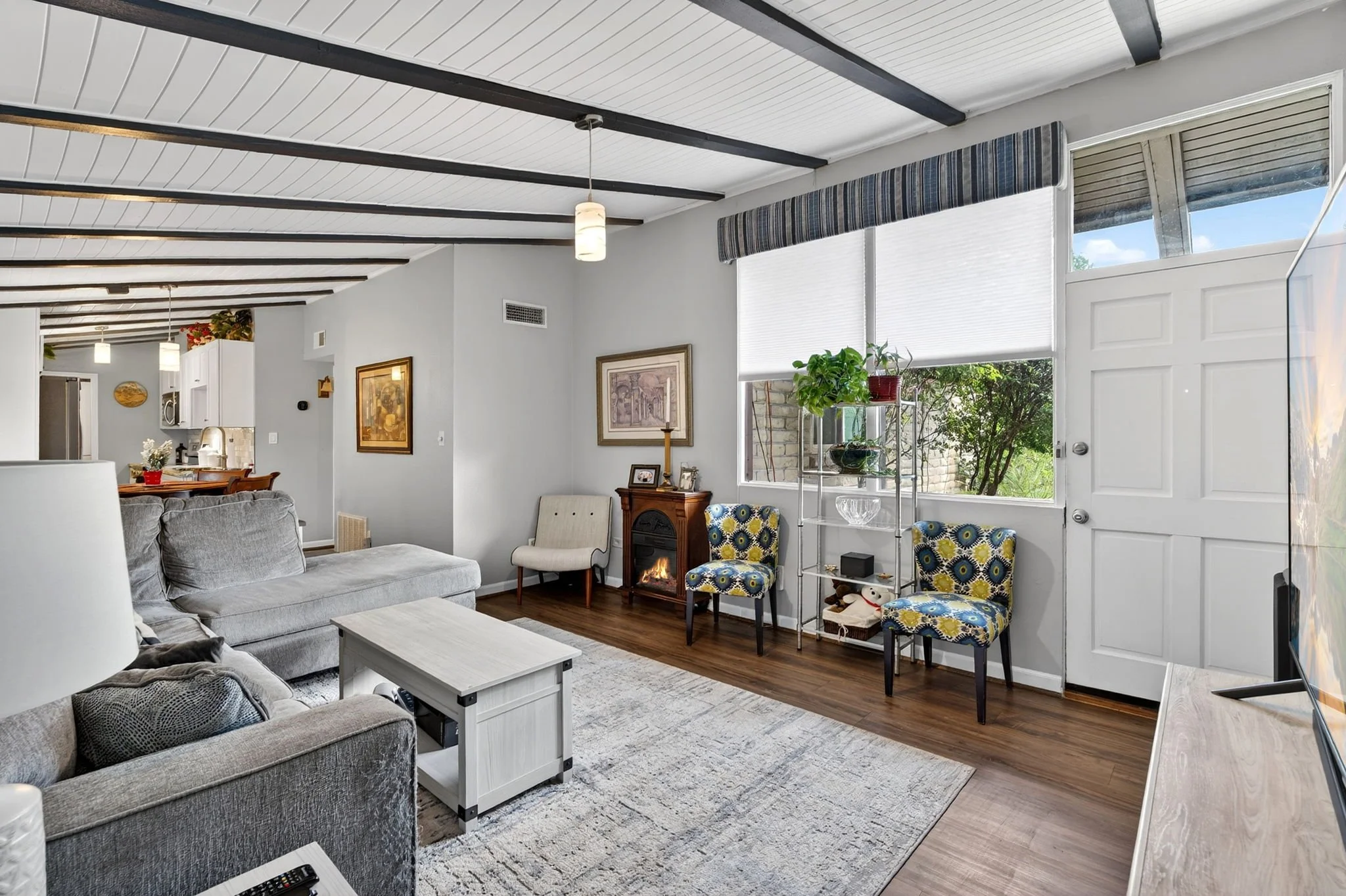 Living room with gray sofa, white coffee table, patterned chairs, small fireplace, wall art, plants, and large windows with blinds, ceiling beams, and pendant lights.