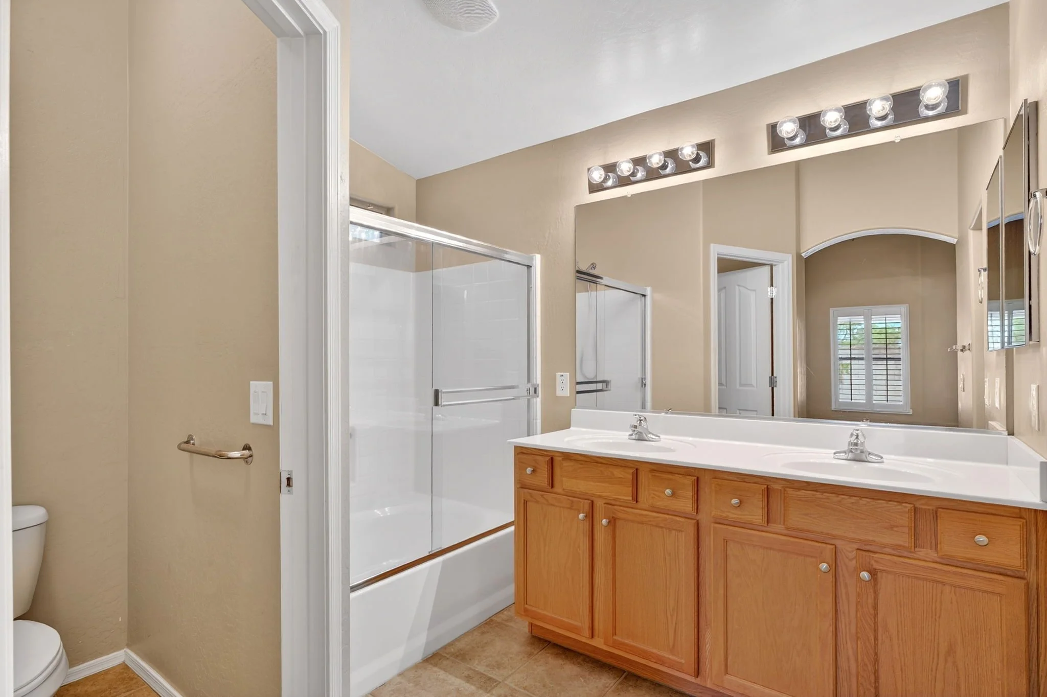 Bathroom with beige walls, two sinks with wooden cabinets, large mirror, and vanity lights. Shower with sliding door, window with blinds, and a toilet partially visible.