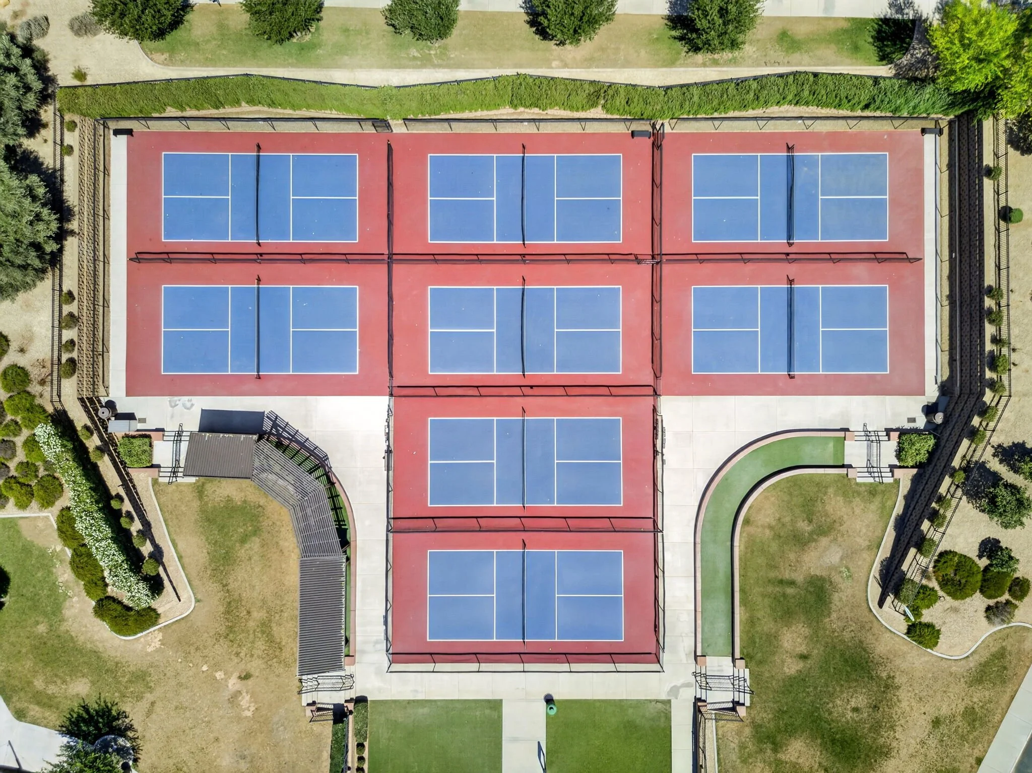 Aerial view of multiple blue tennis courts with white lines, surrounded by green fencing, pathways, and landscaped areas with trees and grass.