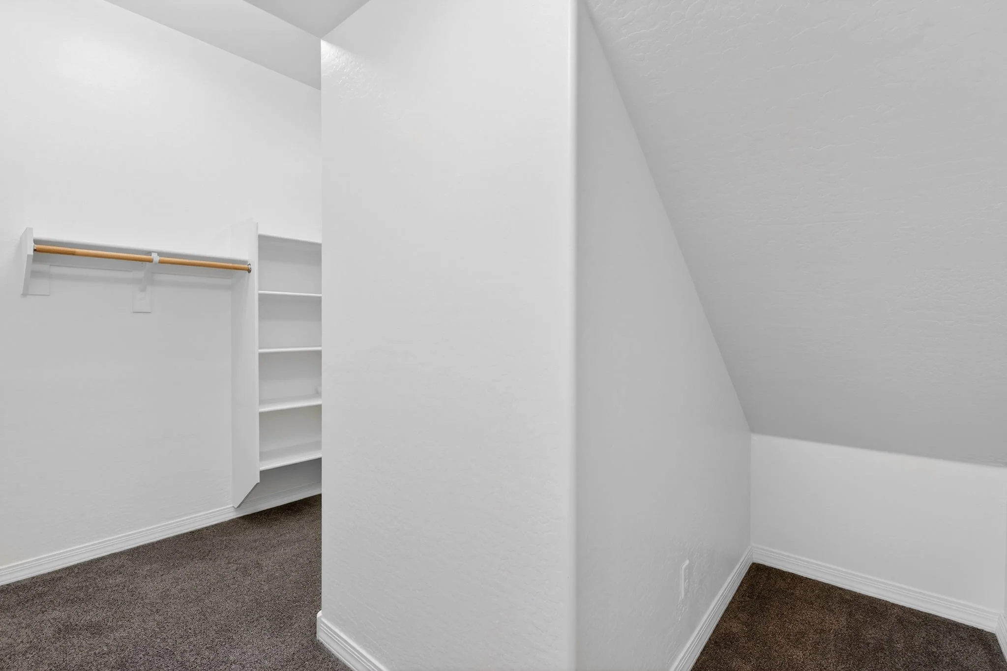 Empty walk-in closet with white walls, a wooden rod for hanging clothes, white shelves, and brown carpeted floor.