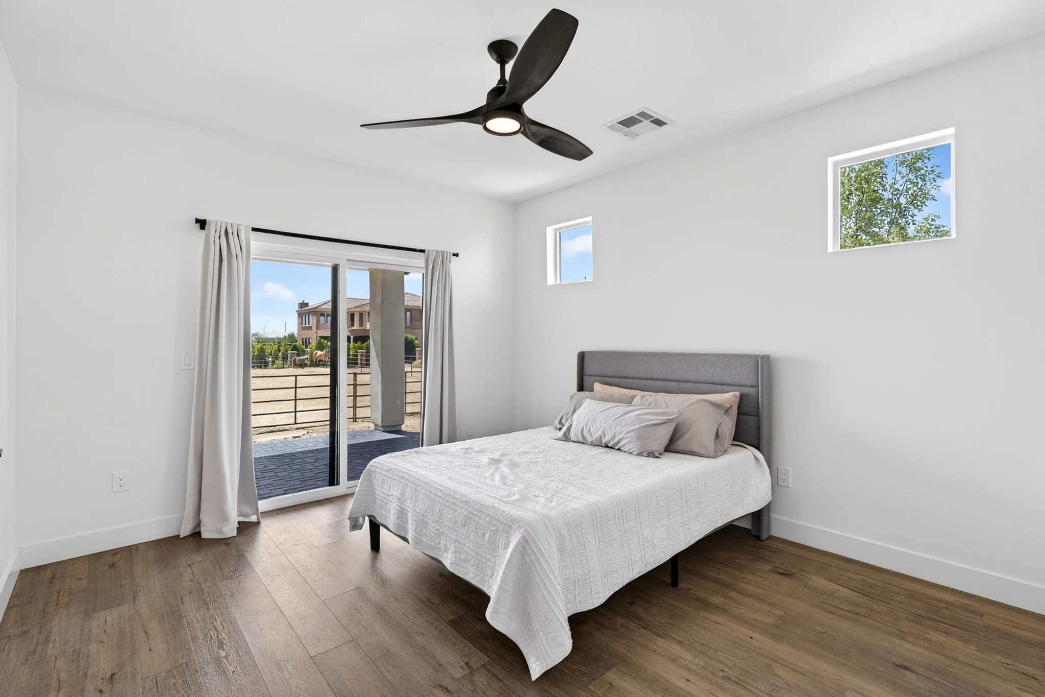 Clean bedroom with wooden flooring, bed with white bedding, gray upholstered headboard, sliding glass door with white curtains, and two small windows showing blue sky and trees.