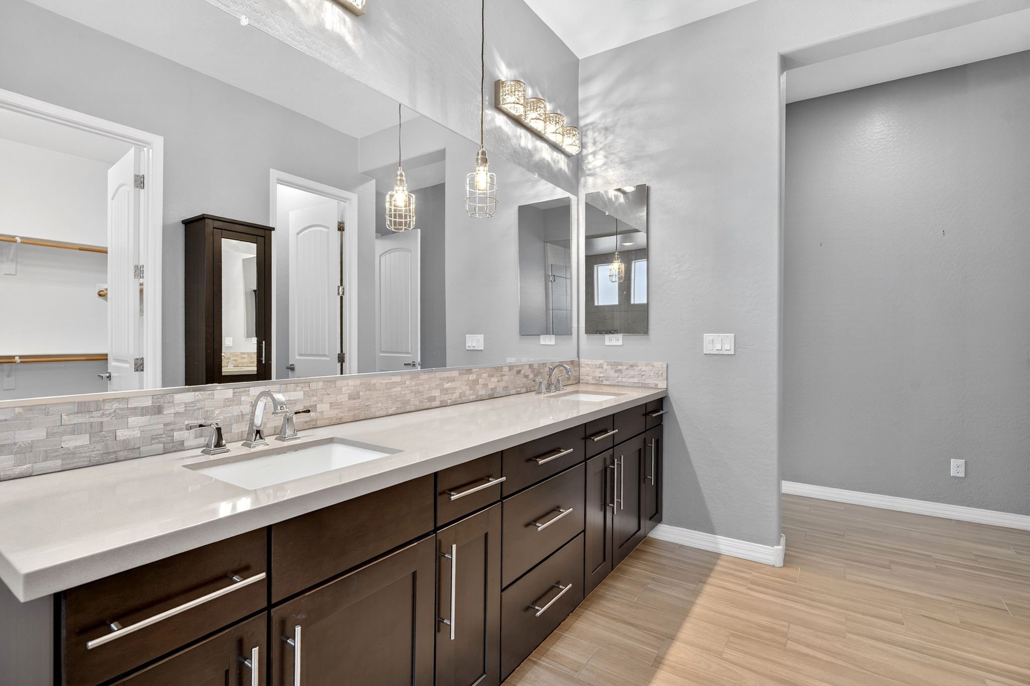 A modern bathroom with a large mirror above a white countertop featuring two sinks and dark wood cabinets. There are hanging light fixtures and a tiled backsplash. A doorway leads to a walk-in closet with wooden shelves.