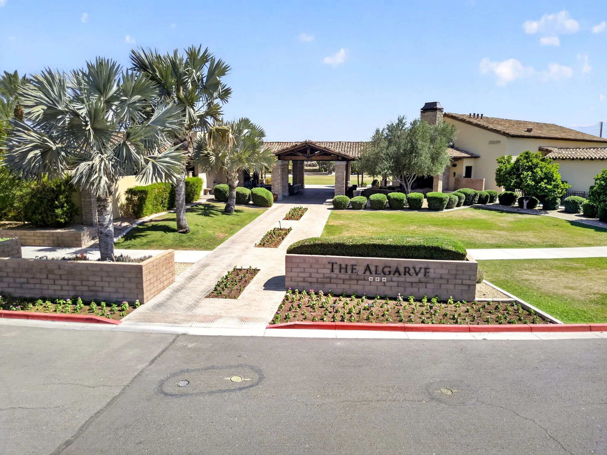 The image shows the entrance to a residential complex called The Algarve, with a well-maintained garden, trees, and a small pavilion or shelter. There are brick planters and paving stones, and the background features a house with a tiled roof.