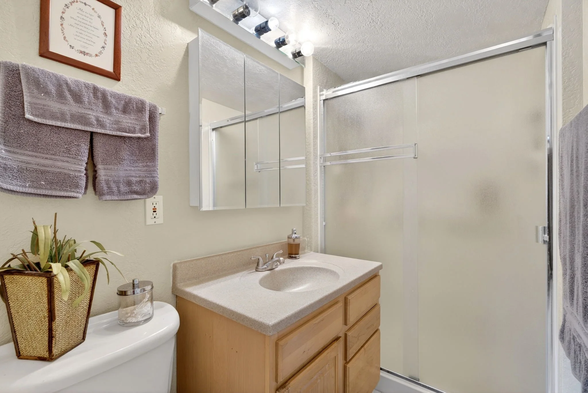 Bathroom with beige countertop, wooden cabinet, silver faucet, 2 purple towels on towel rack, framed picture on wall, small plant, mirror, soap dispenser, and glass of water.