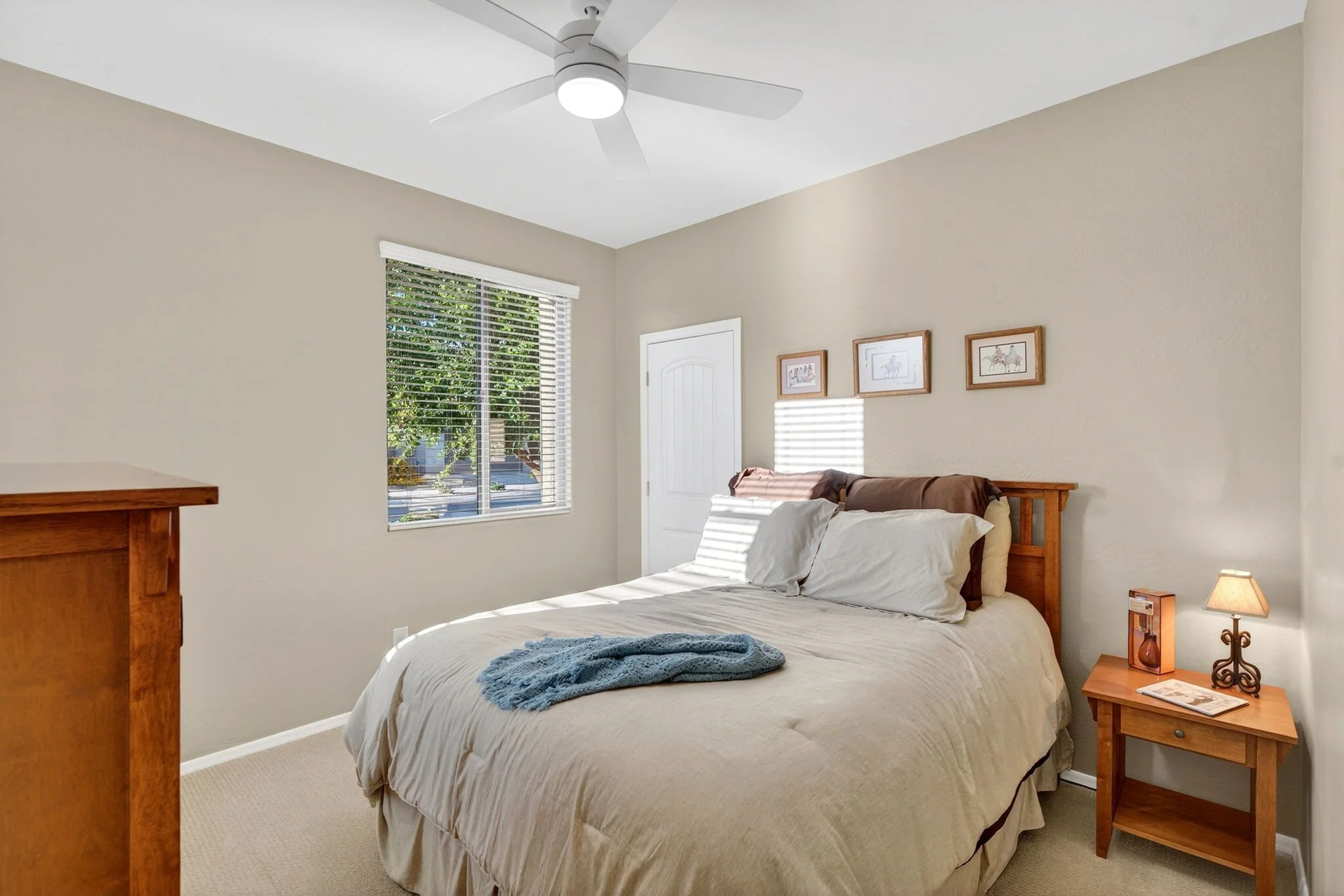 A bedroom with a bed, wooden nightstand, window with blinds, ceiling fan, and framed pictures on the wall.