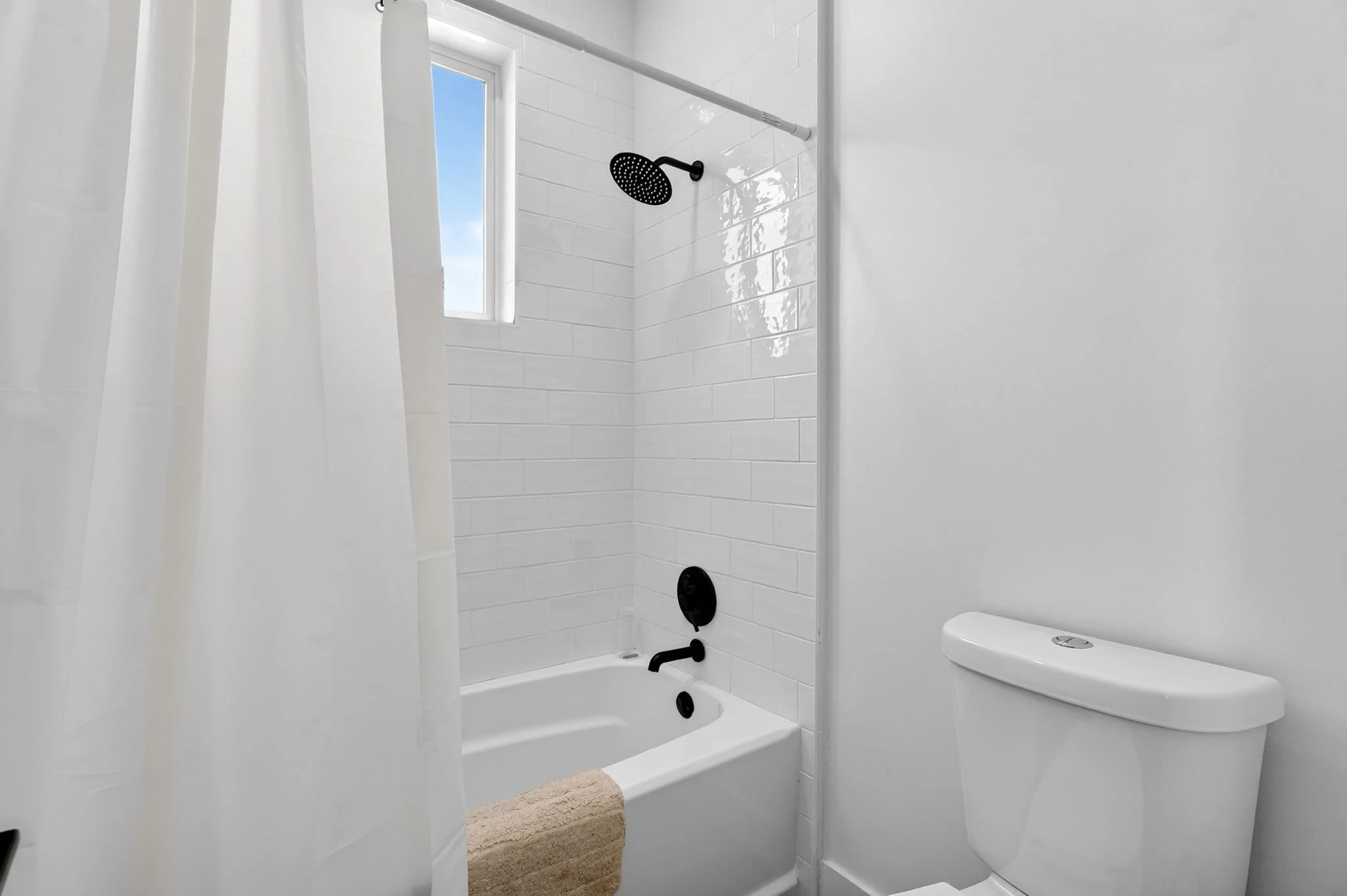 Small white bathroom with bathtub, shower with black fixtures, white tile walls, small window, and toilet, with a beige bath mat.