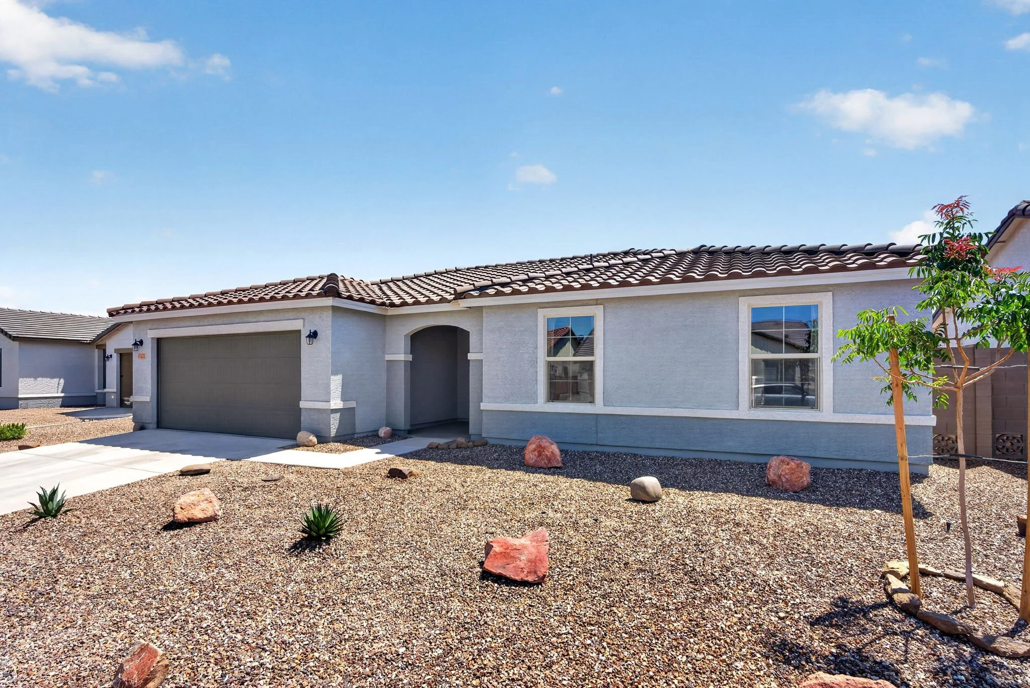 Front view of a single-story house with a gray garage door, white stucco walls, tiled roof, and two windows, with desert landscaping including rocks, small plants, and a young tree under a blue sky with scattered clouds.