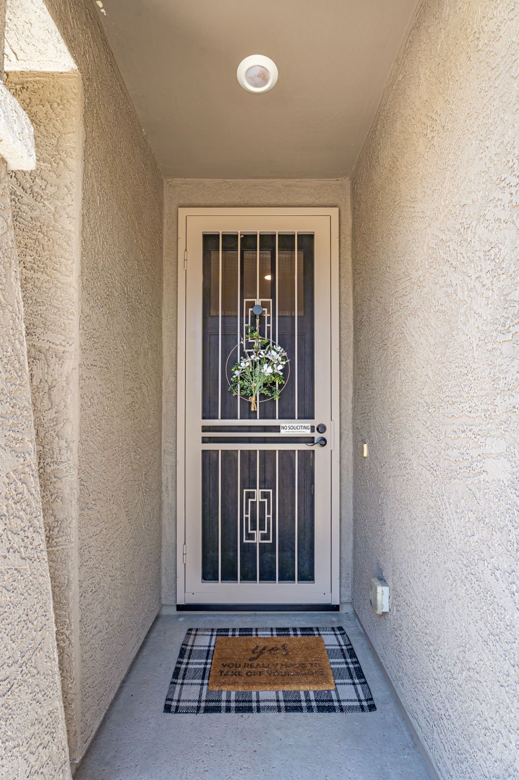 Front door with a decorative wreath, black door behind a security screen, and a 'No Soliciting' sign, on a small porch with a plaid doormat and beige textured walls.