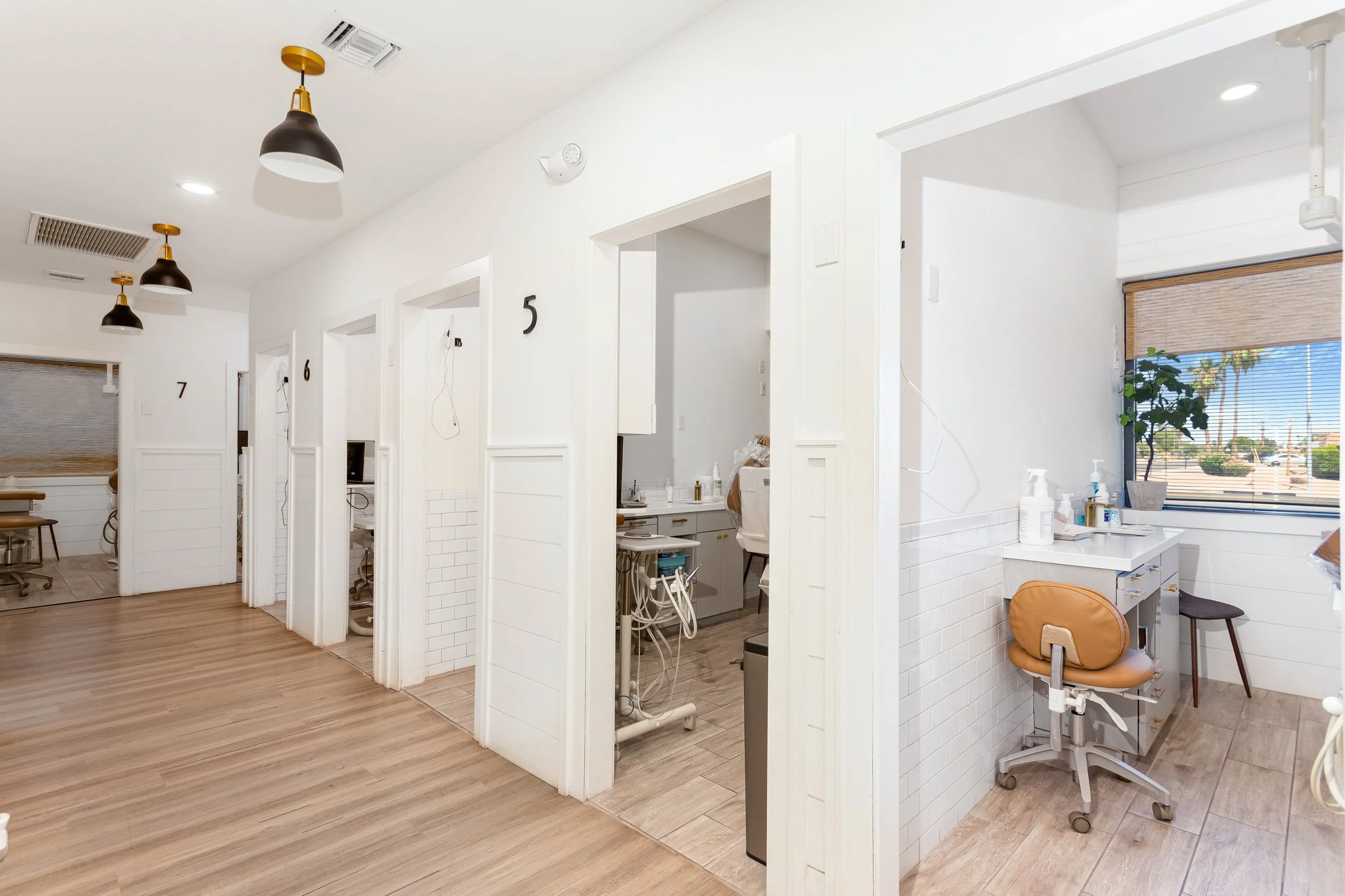 Interior view of a modern medical or dental clinic with white walls, numbered treatment rooms, lighting fixtures, window with blinds, and dental chairs visible in some rooms.