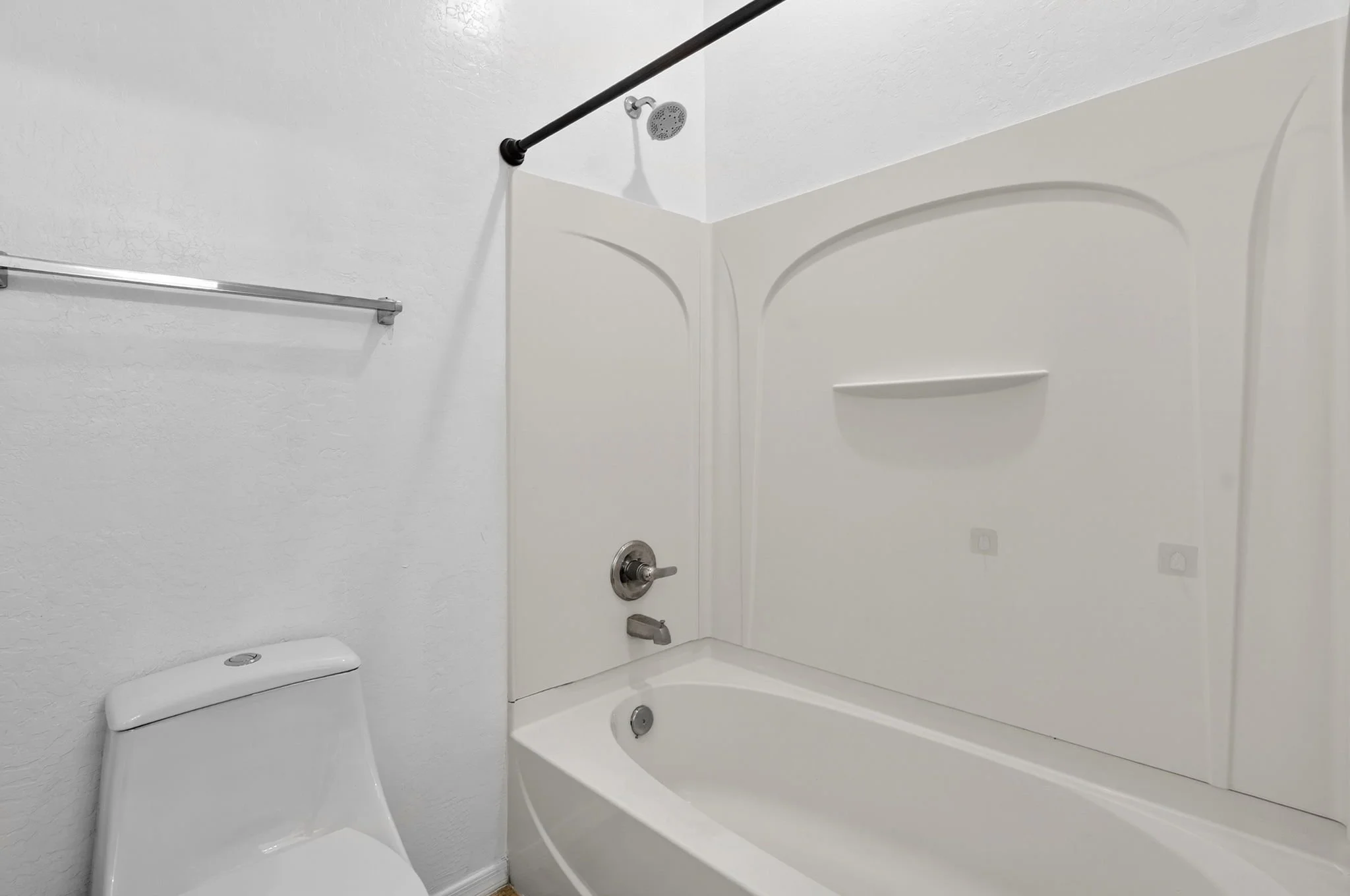 A white bathroom shower area with a showerhead, a grab bar, and a pinkish-tan wall. The shower has a built-in shelf and a lower drain. To the left, there is a white toilet with a closed lid.