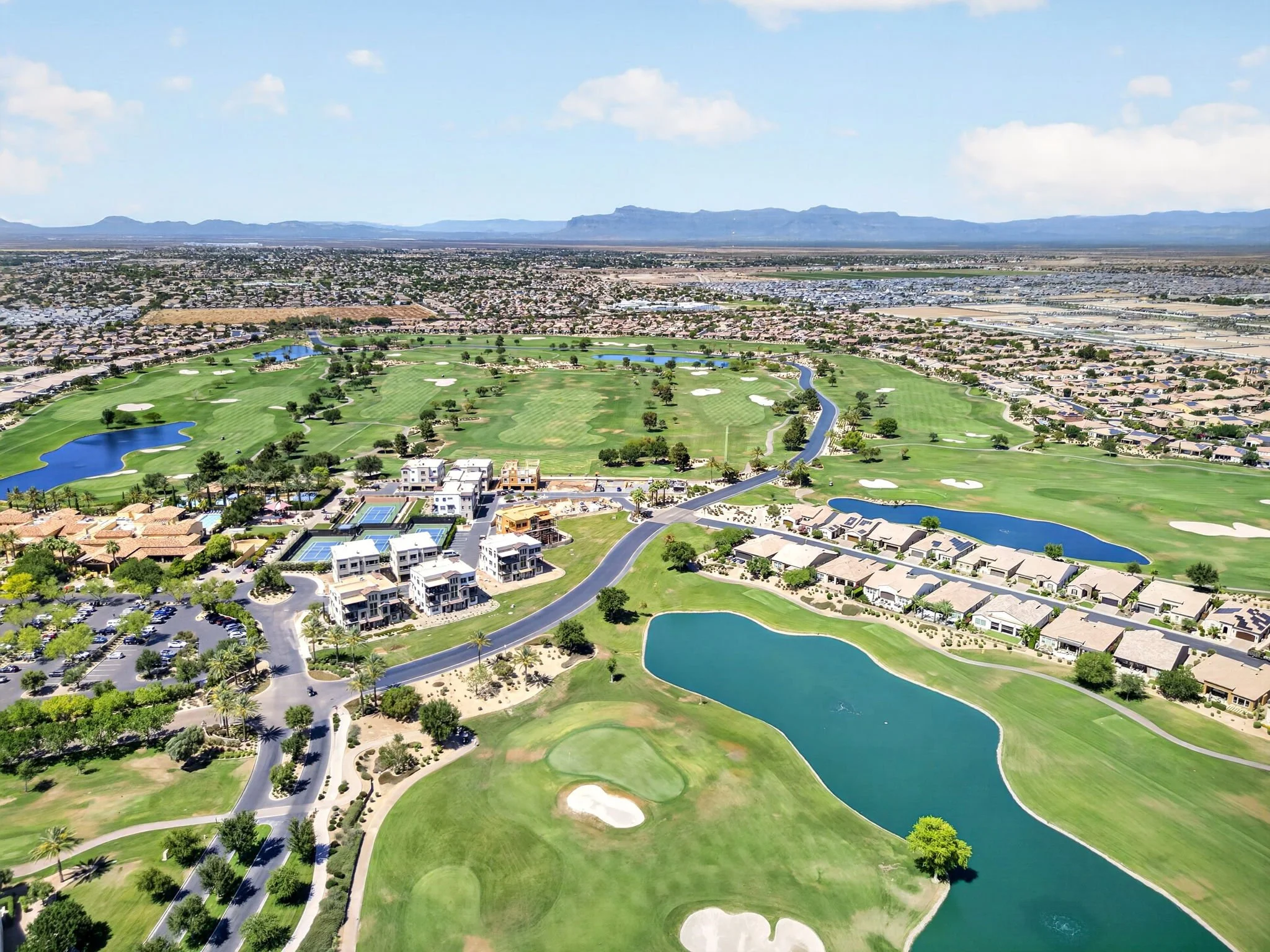Aerial view of a golf course with water hazards, residential buildings, parking lots, and mountains in the distance under a partly cloudy sky.