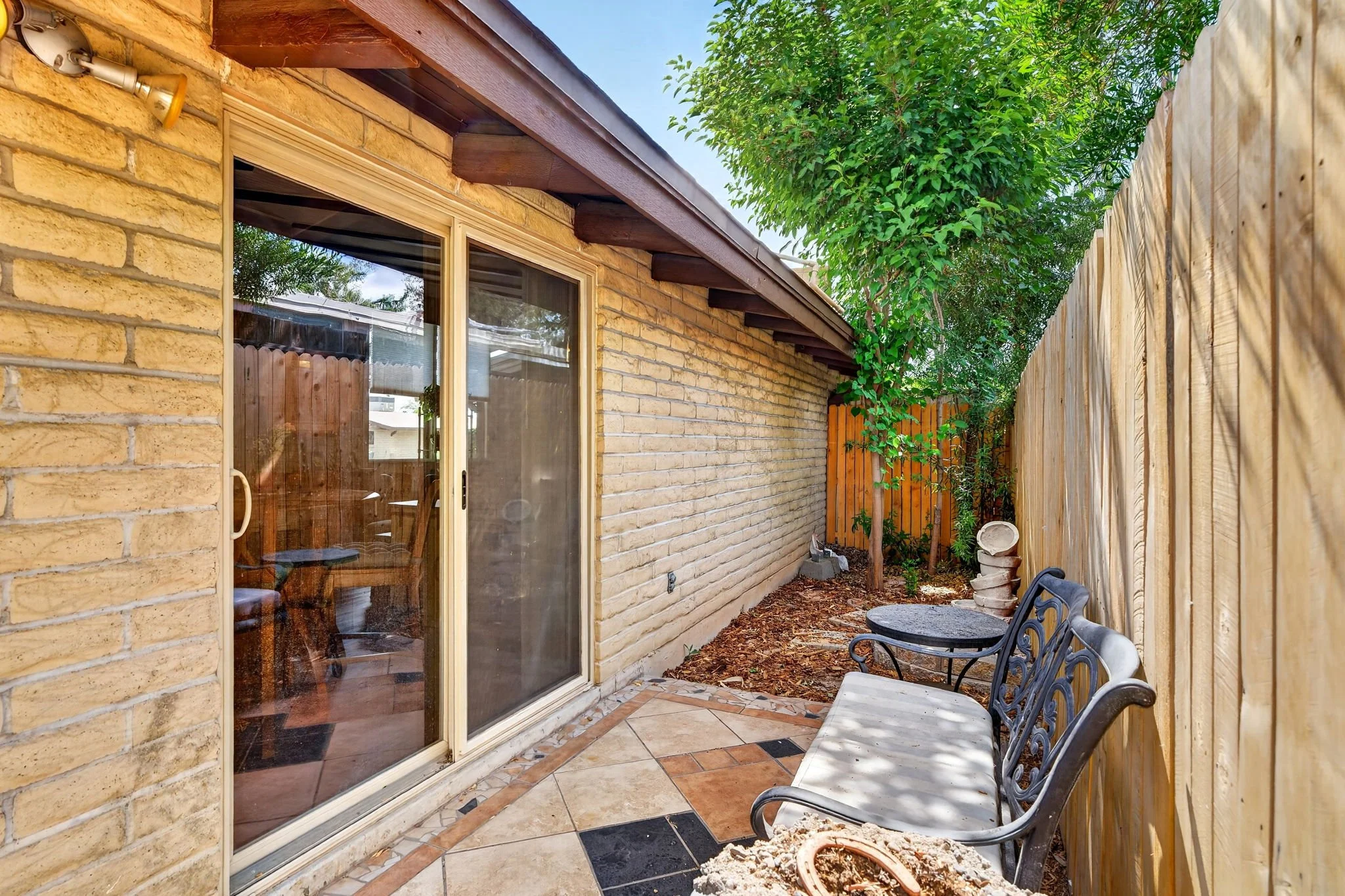 An outdoor patio area next to a brick house with a sliding glass door, fenced by a wooden privacy fence, with chairs and a potted plant visible.