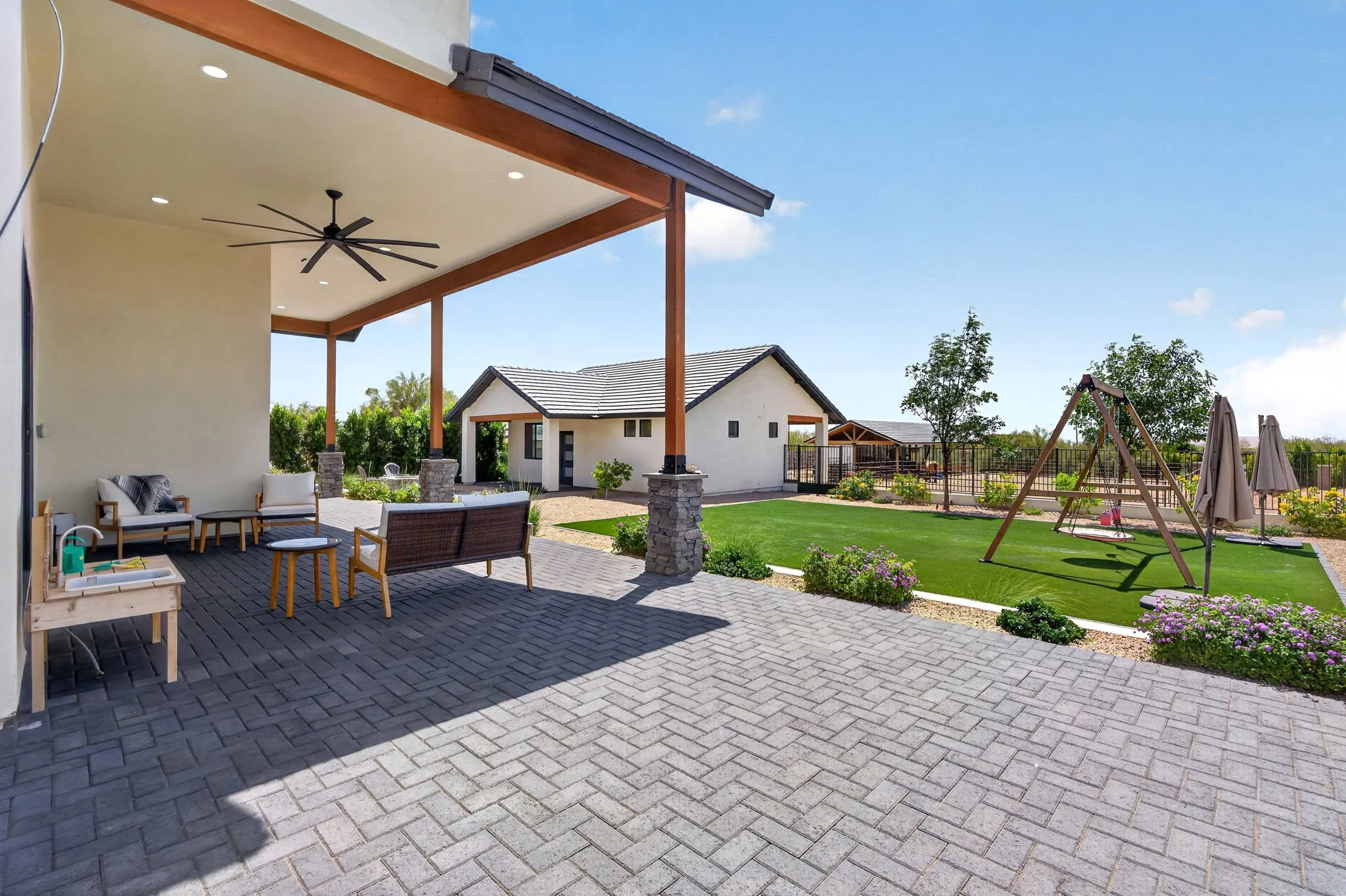 Backyard patio with outdoor seating, a covered porch, a grass lawn, a children's swing set, a small garden with purple flowers, and neighboring houses under a blue sky.
