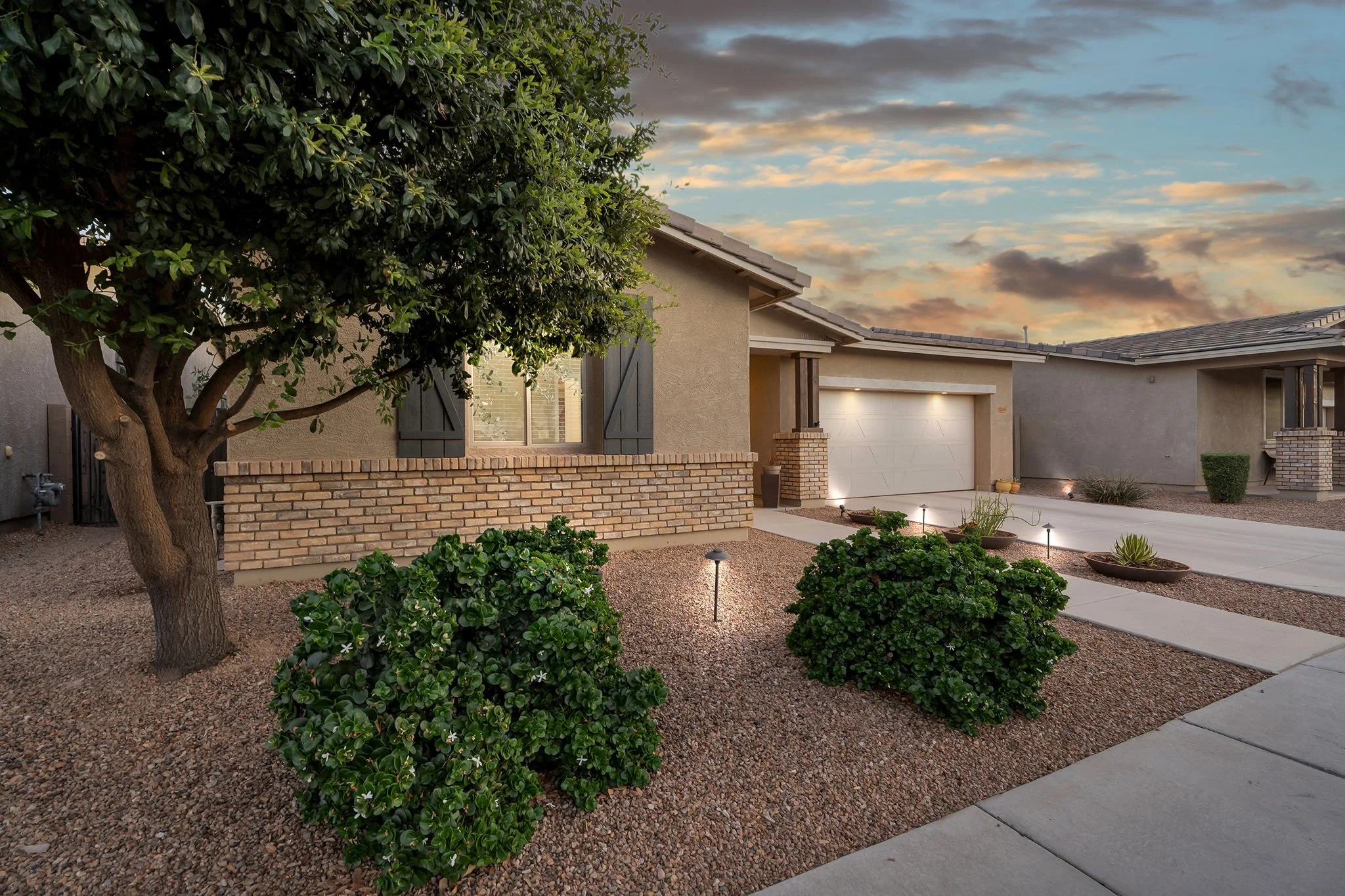 A suburban house with a brick wall and a large tree in the front yard during sunset. The scene includes a driveway, small shrubs, and garden lighting.