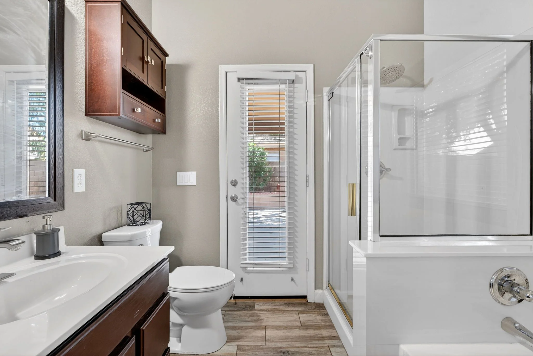 A bathroom with a white toilet, dark wood vanity, white sink, mirror, and shower with glass doors. A window with blinds and a door with blinds are visible.