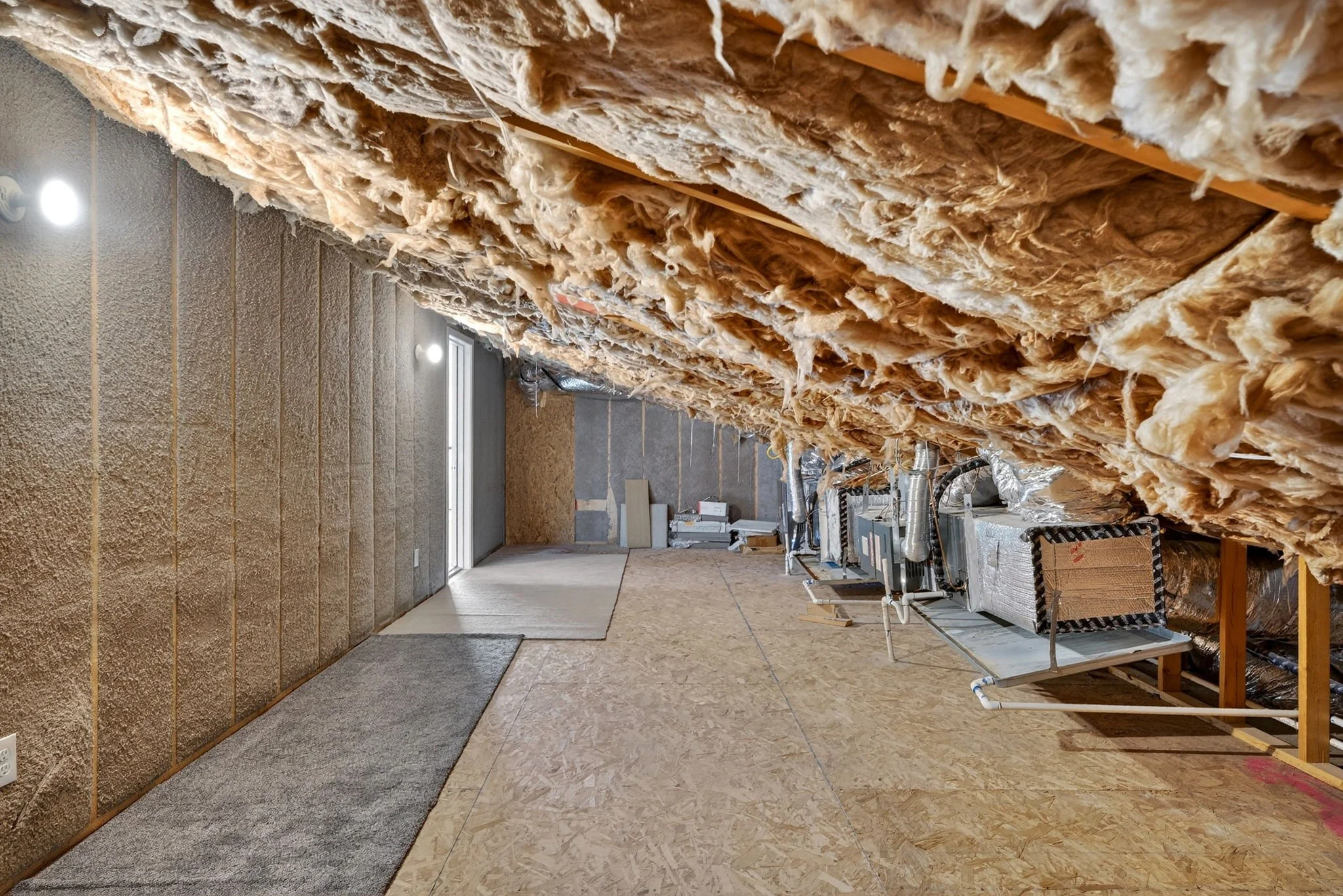 Construction site showing unfinished interior with insulation installed in ceiling, exposed beams, and incomplete walls. Fluorescent lights are mounted on the ceiling.