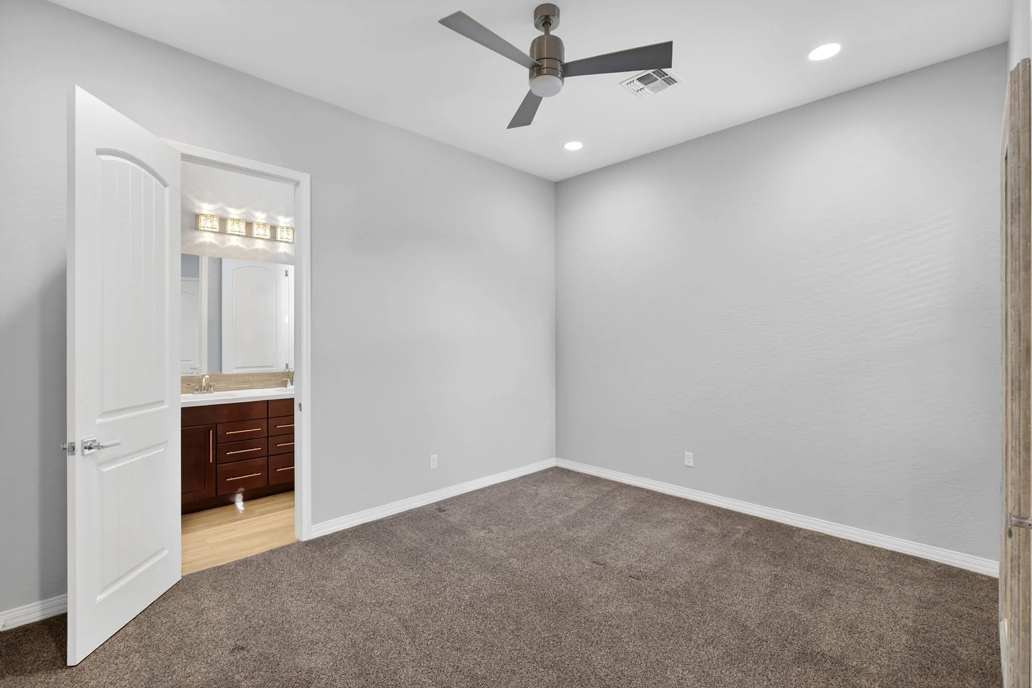 Empty room with gray walls, carpeted floor, ceiling fan, and a doorway leading to a bathroom with wood vanity and mirror.