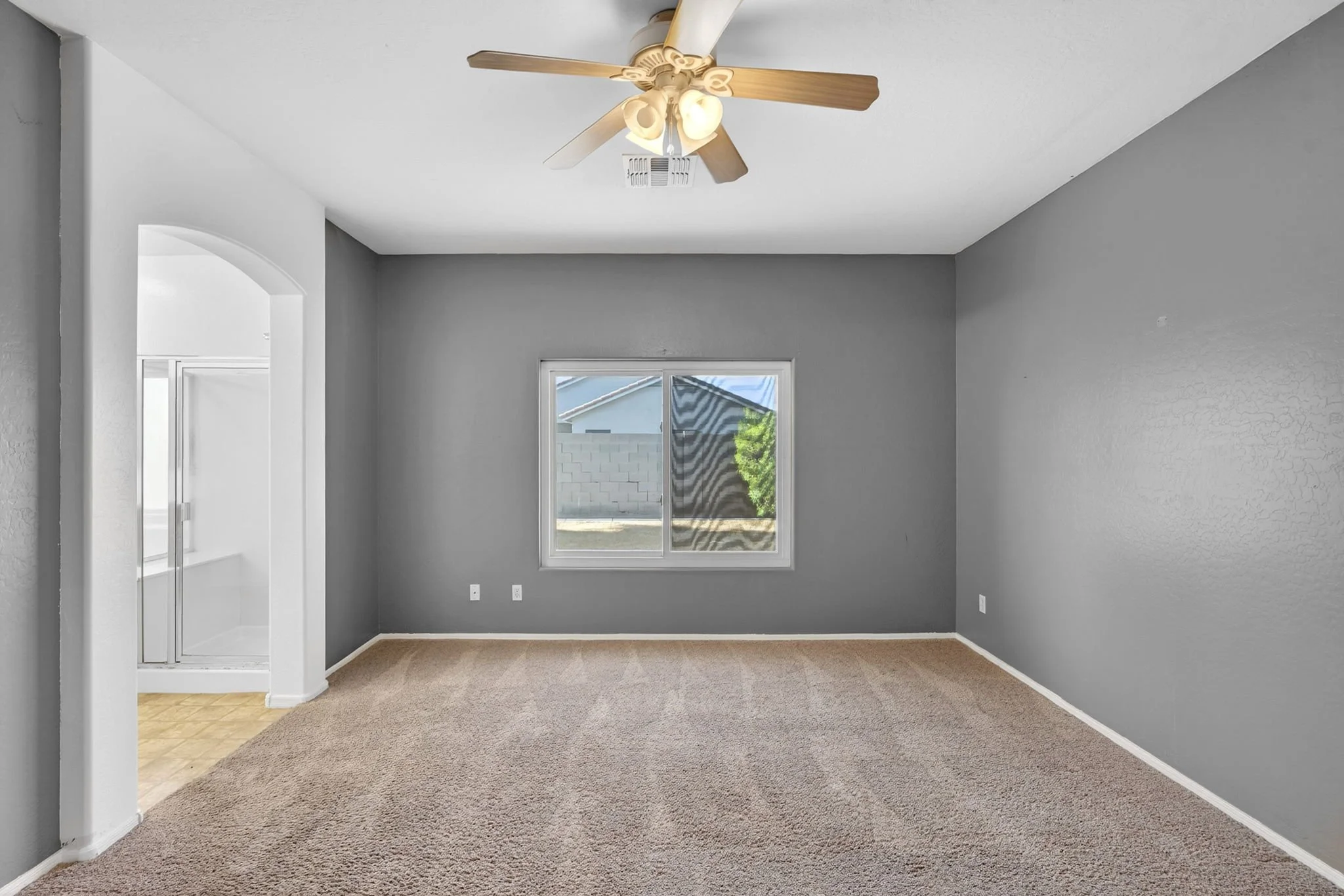 Empty living room with gray walls, a window, a ceiling fan with four wooden blades and light fixtures, and beige carpeting.