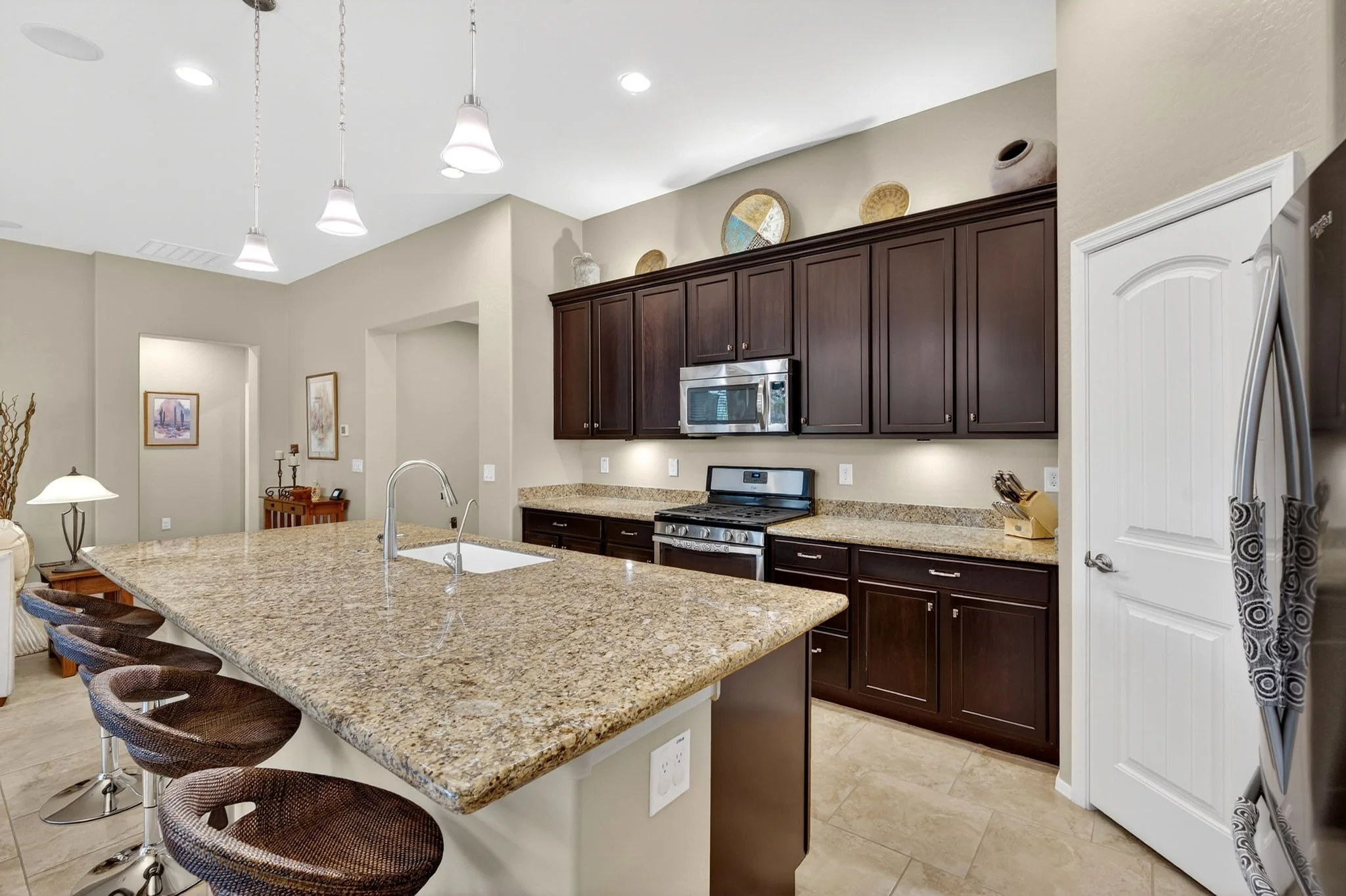 Kitchen with granite island, dark wood cabinets, stainless steel microwave and stove, beige walls, and pendant and recessed lighting.
