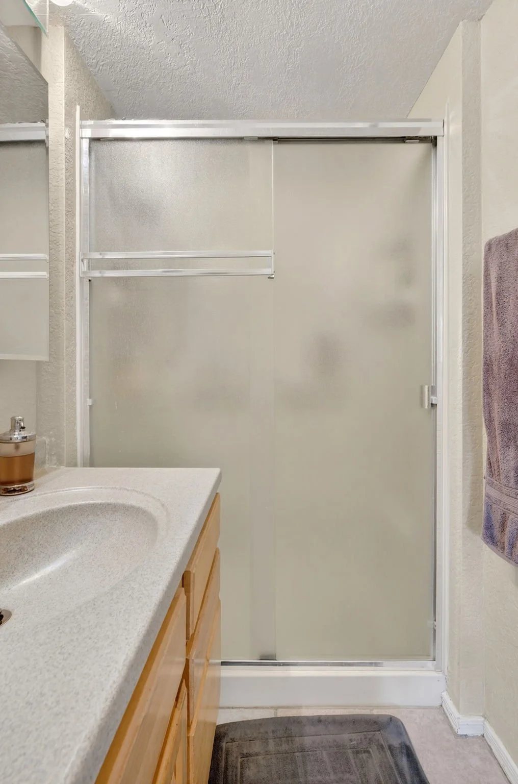 A bathroom with a frosted glass shower door, a beige countertop with an integrated sink, a wooden cabinet below, a purple towel hanging on the wall, and a gray rug on the floor.