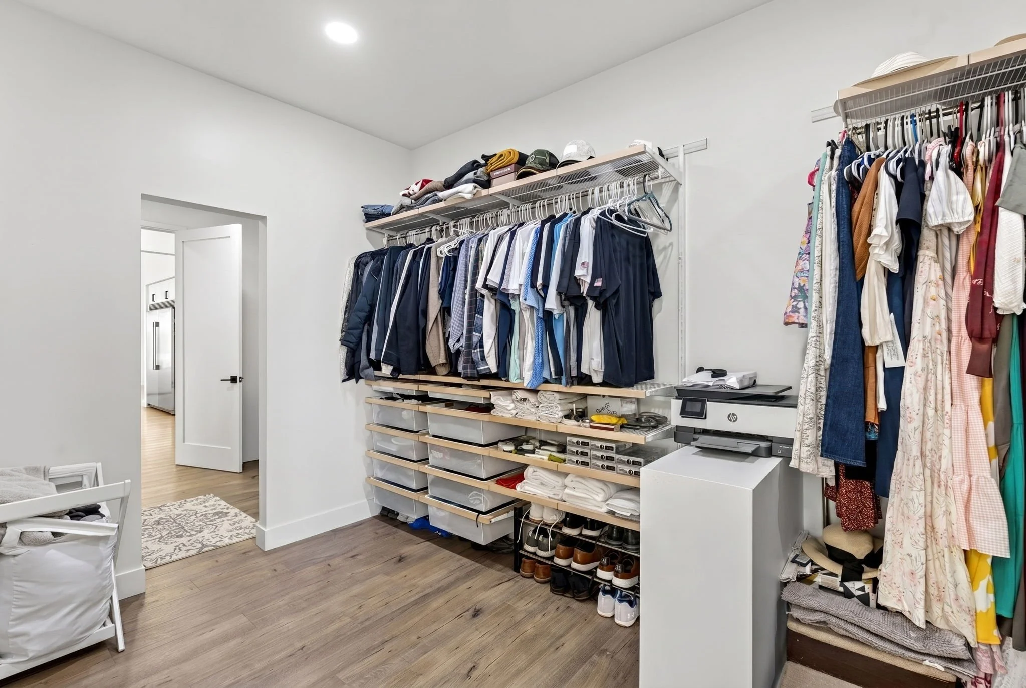 A walk-in closet with white walls and wooden flooring, featuring organized clothing and shoes on shelves and racks, a desk with a printer, and a doorway leading to another room.