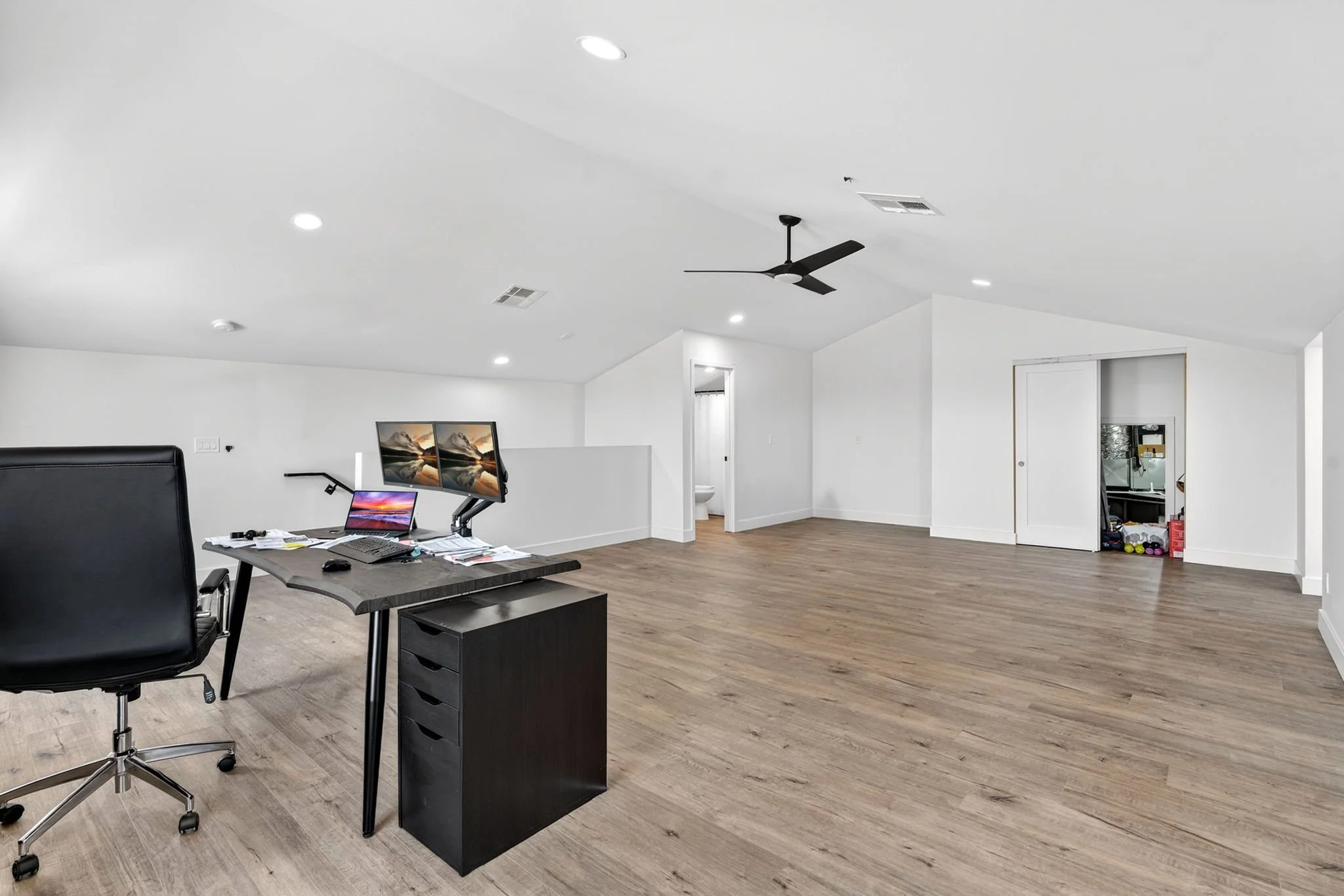 An empty modern office space with a desk, computer monitors, a black office chair, and hardwood flooring. The ceiling has recessed lighting, a ceiling fan, and vent. The walls are white, with a small room or closet visible in the background with exer