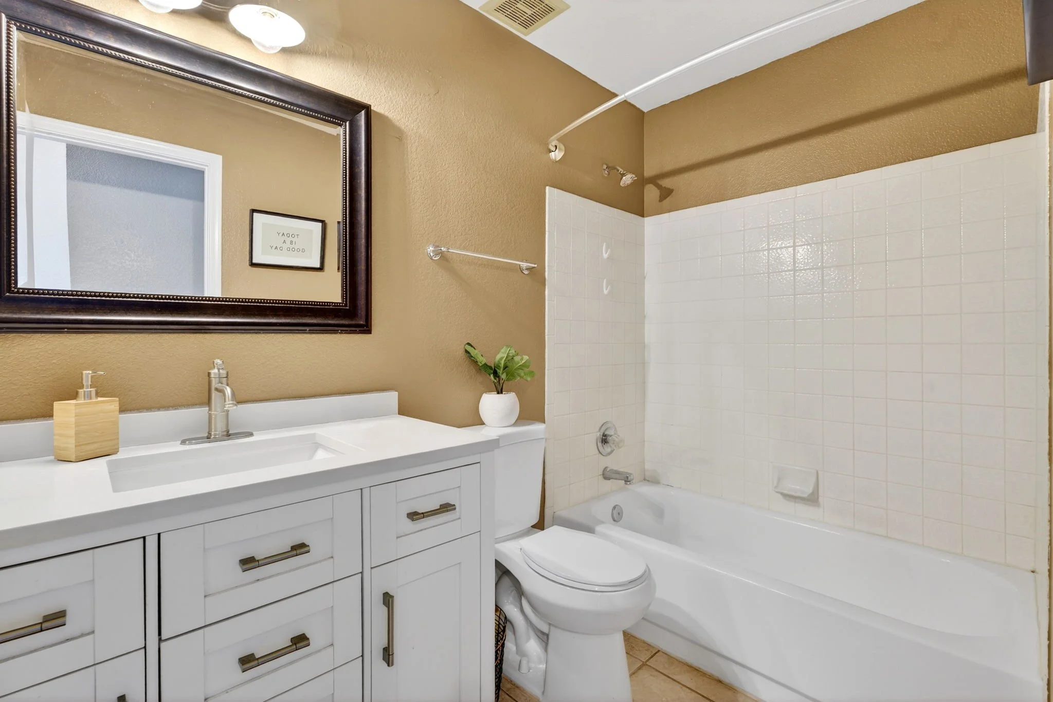 Bathroom with a vanity mirror, white vanity with drawers and gold handles, beige walls, framed picture, plant, bathtub with shower curtain, and toilet.
