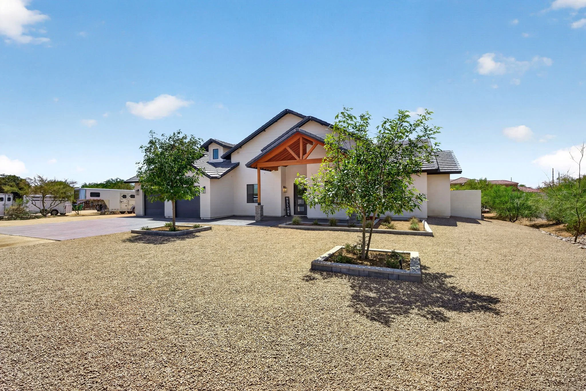 A modern house with a light-colored exterior, dark roof, front porch, and landscaped yard with small trees, surrounded by a gravel driveway and a clear blue sky.
