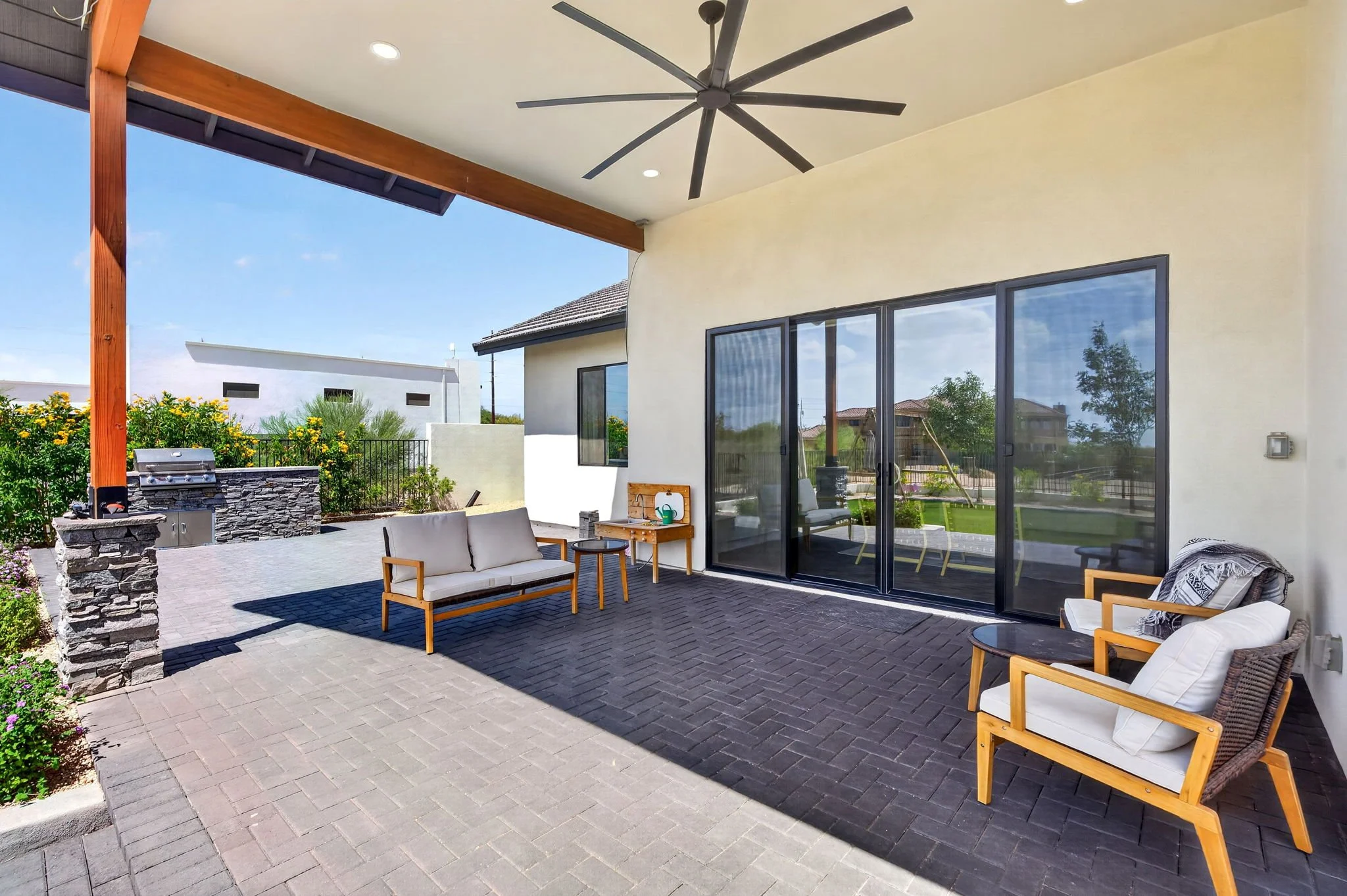 A covered patio area with outdoor furniture including a sofa, chairs, and a small table. There is a ceiling fan above and sliding glass doors leading to the interior of the house. In the background, there is a garden with flowering plants, a grill, a