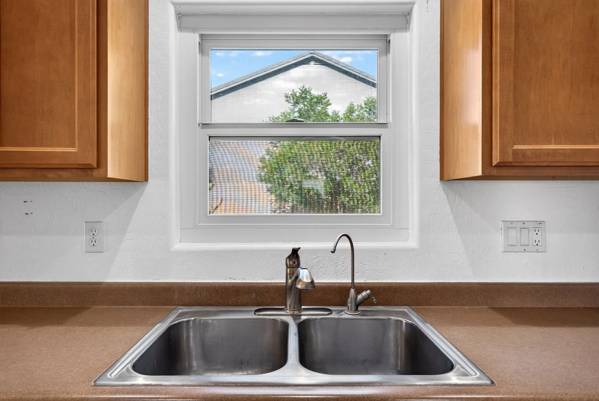 Kitchen sink with faucet beneath a double-pane window showing a house and trees outside.