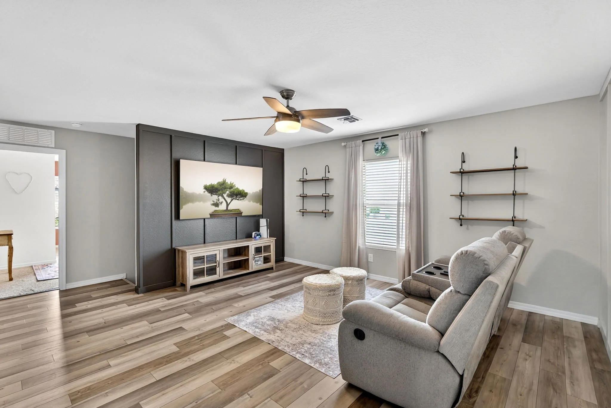 Living room with gray walls, wooden flooring, a ceiling fan, a TV on a TV stand, and beige furniture including a reclining chair and ottomans. There are wall shelves and a window with curtains.