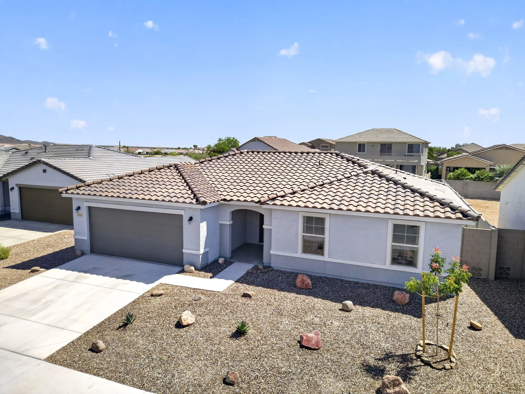 View of a residential neighborhood with a focus on the front yard of a house that has a white exterior, gray garage doors, and a tiled roof. The yard is landscaped with gravel, rocks, small plants, and a young tree, under a bright blue sky with a few
