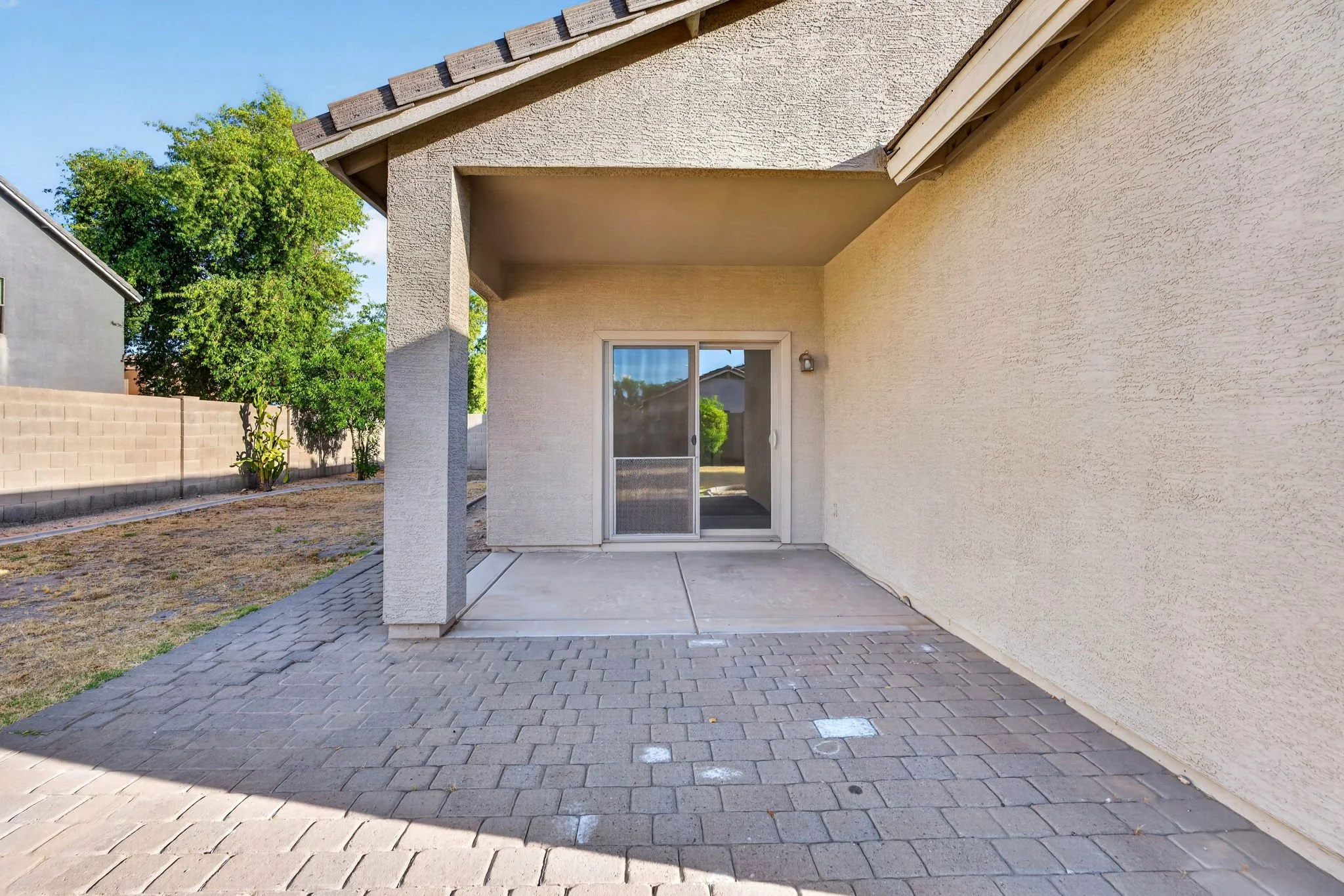 View of a backyard patio area with concrete flooring, sliding glass door, and beige stucco wall. The patio is shaded by the house's roofline, and there is a small garden bed with green plants along the fence. The yard is enclosed by a beige block fen