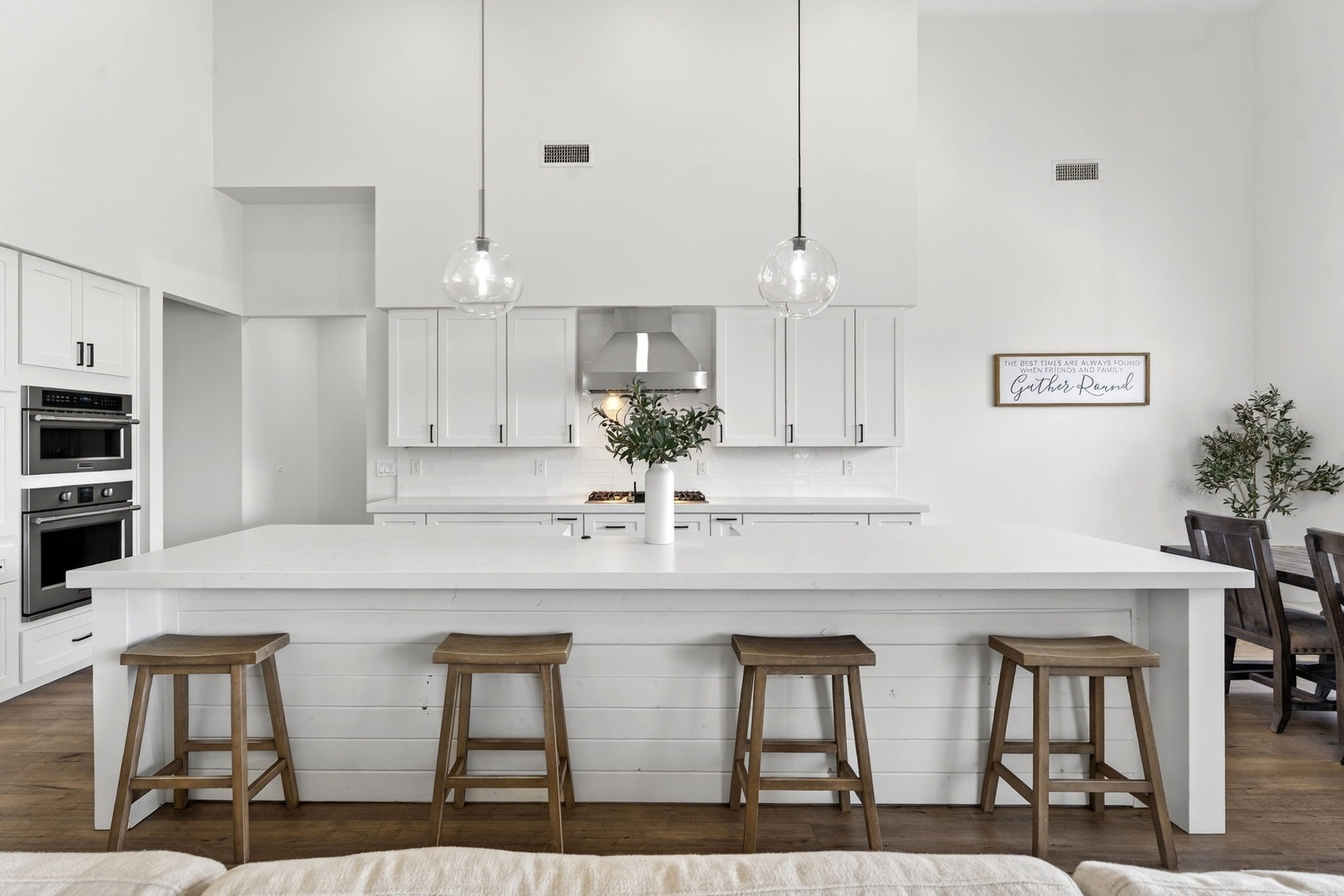 Modern kitchen with white cabinets, a white island with a vase and greenery, stainless steel appliances, wooden stools, black chairs, a wall sign, and green plants.