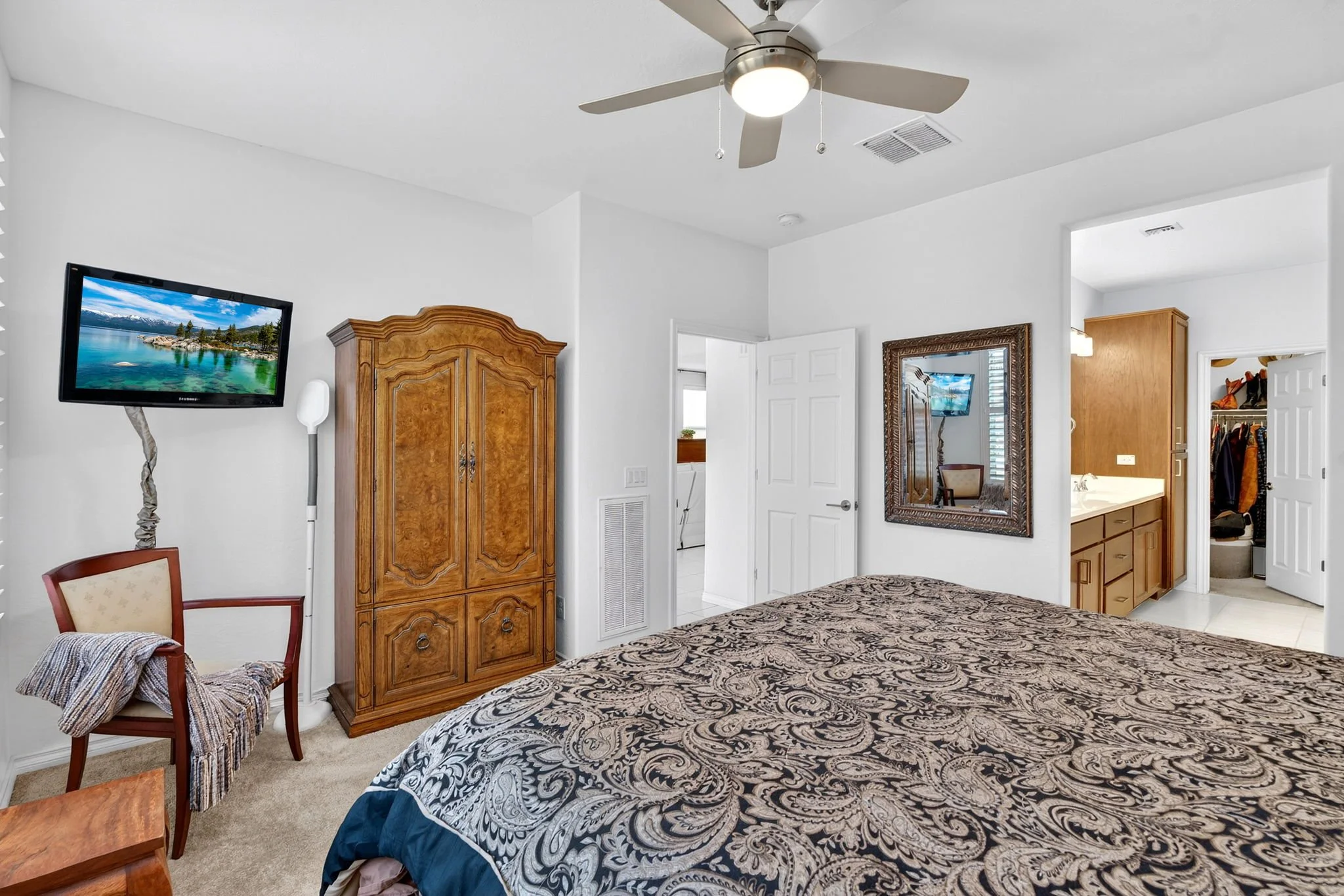 A bedroom featuring a patterned bedspread, a wooden armoire, a wall-mounted TV, a mirror, and a ceiling fan with light, with an open door leading to a closet.